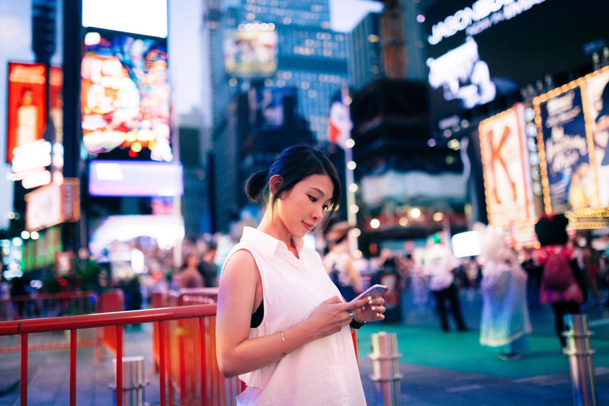 A woman in a white sleeveless top stands in New York City's Times Square at dusk, looking down at the smartphone in her hands