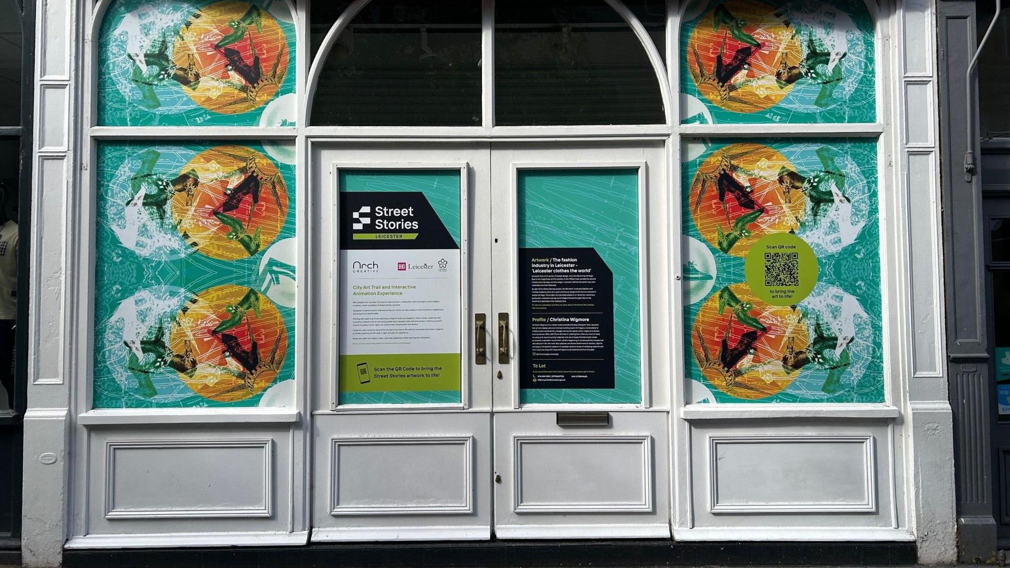 A blue based artwork with colourful birds painted onto the windows of a shopfront, which is predominantly white.