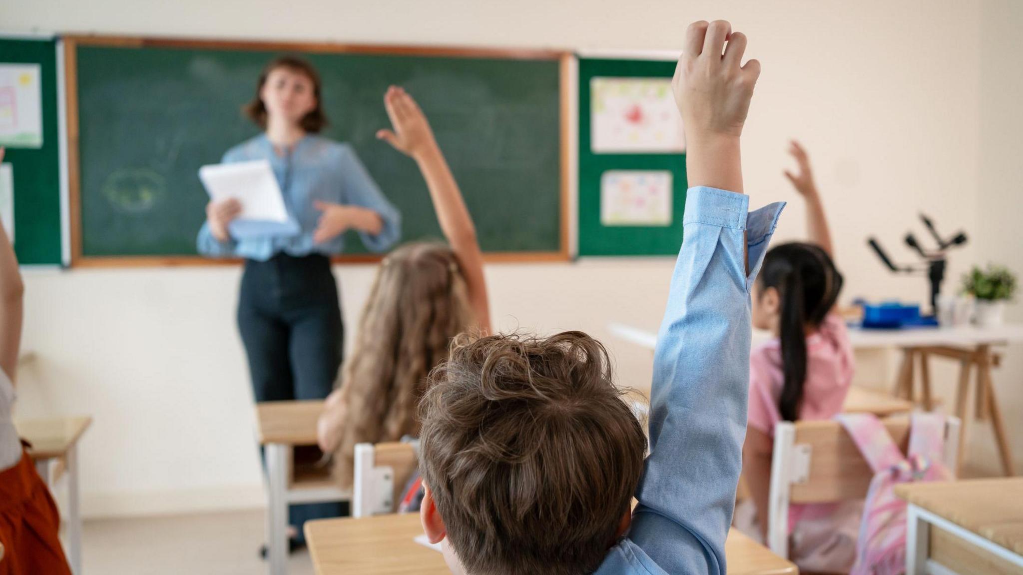 Stock image of youngsters in a classroom. The pupils are seen from behind and several have raised their right arm, while a female teacher stands at the front of the class.