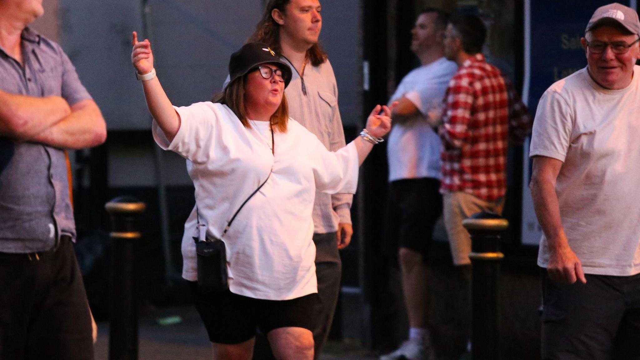 A woman in a black bucket hat and a white T-shirt holds her arms wide as she sings in the street surrounded by other people after Oasis at Heaton Park