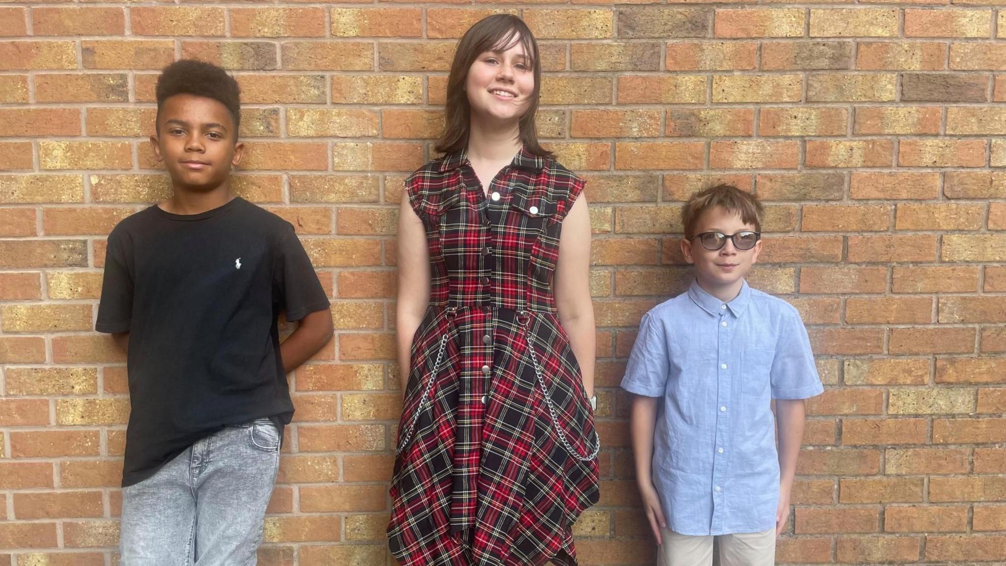 Two teenage boys and a teenage girl line up against a brick wall in their best clothes