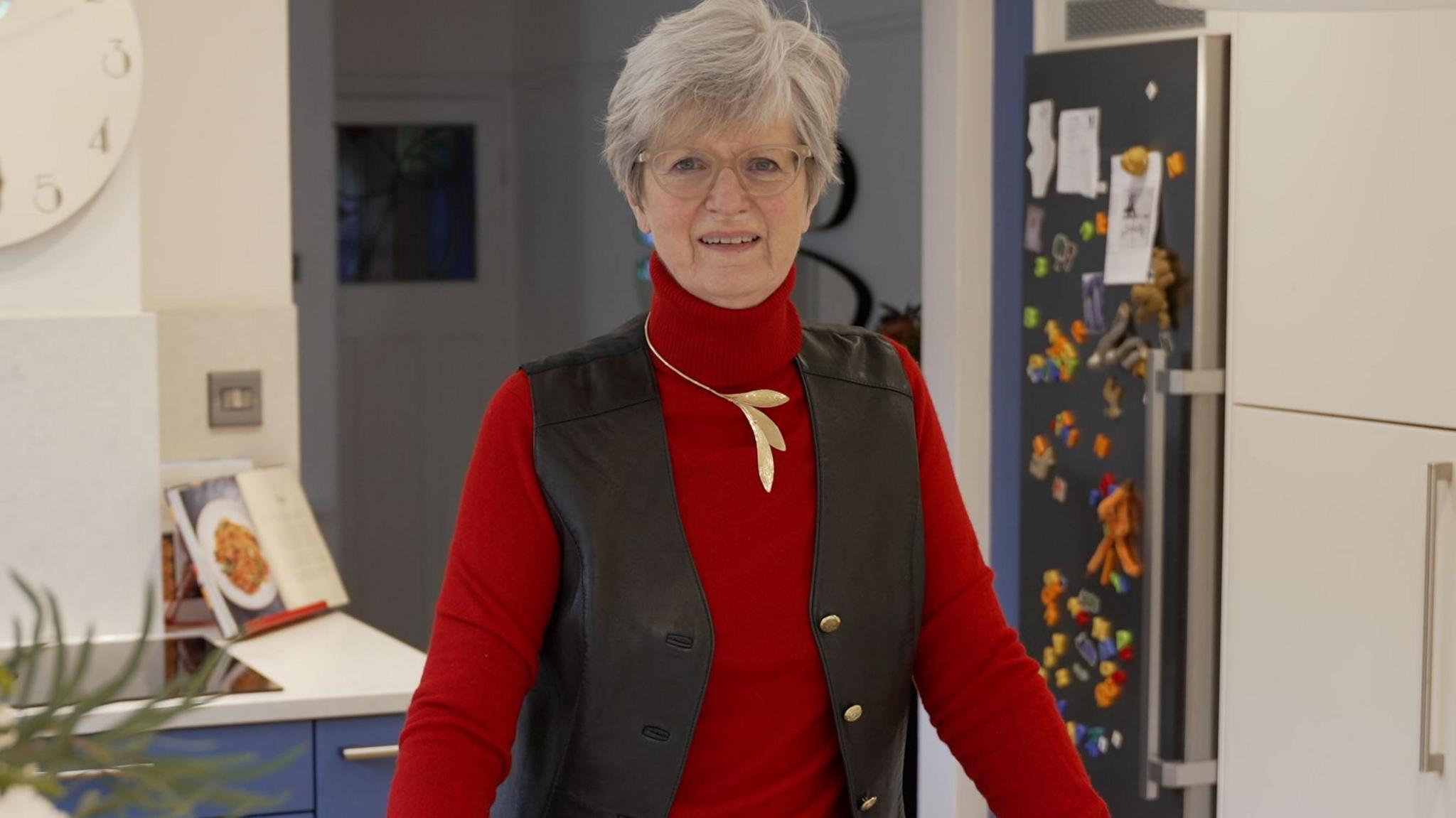 Judith Leary-Joyce standing in her kitchen, looking straight at the camera and smiling. She has short grey hair and is wearing glasses, a red jumper, gold leaf necklace and brown jacket. She has a fridge full of magnets on it behind her, and a cookbook in the left corner open.