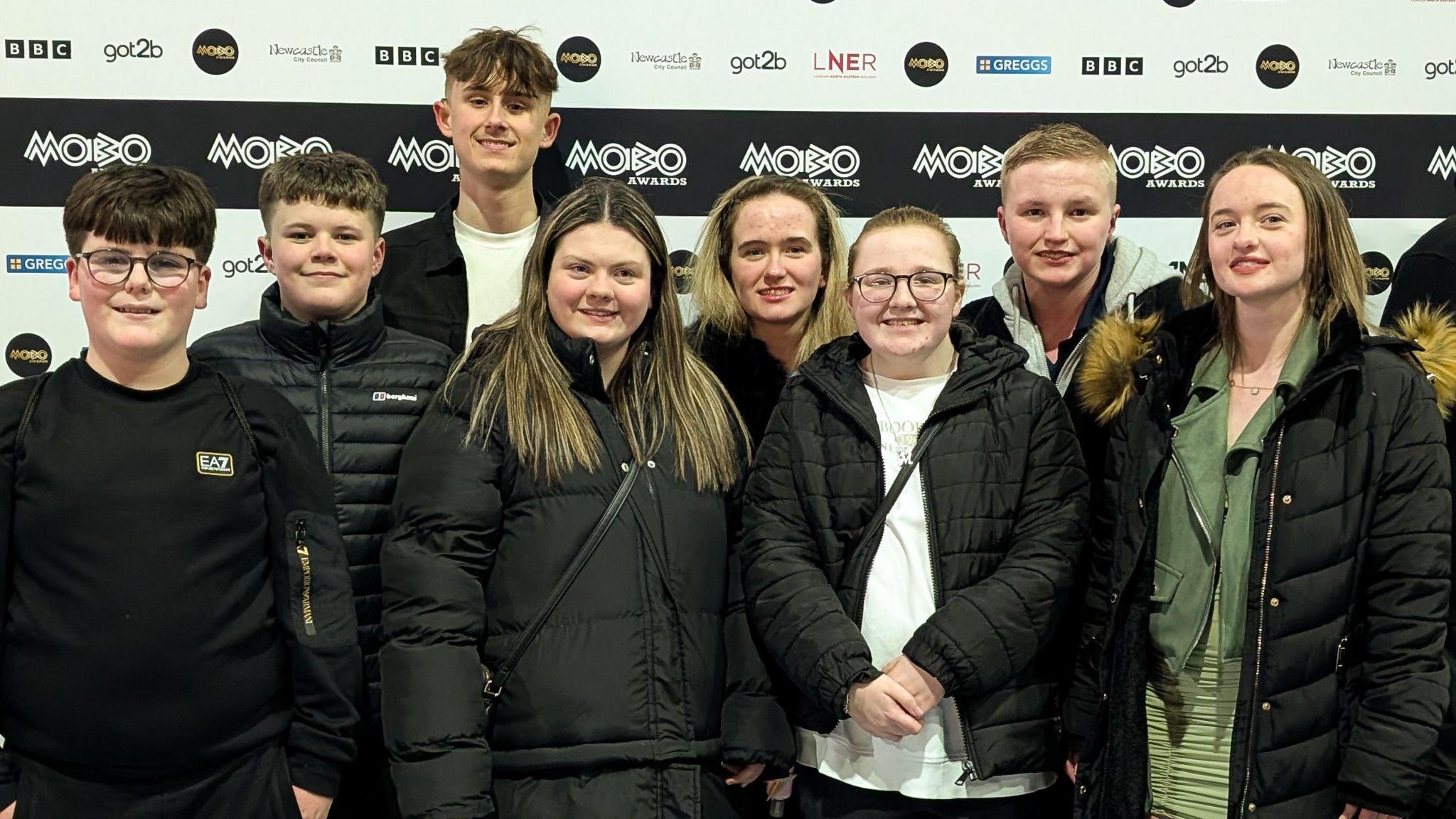 Eight of the 13 pupils who signed on a video at the MOBO Awards. The group of teenagers are all wearing black coats and standing in front of a MOBO banner. 