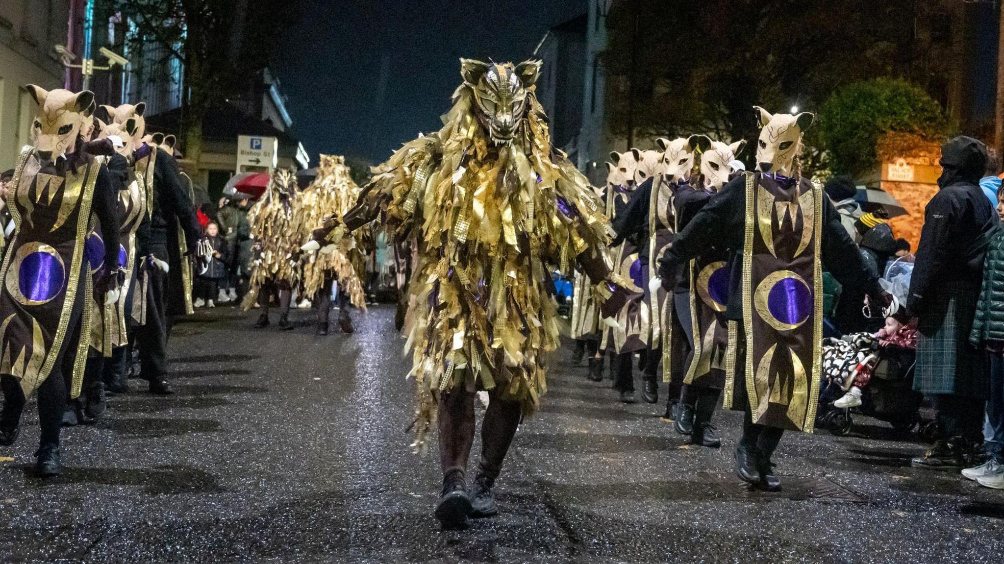 Shows more than a dozen people dressed as wolves in golden and black outfits walking through a street.