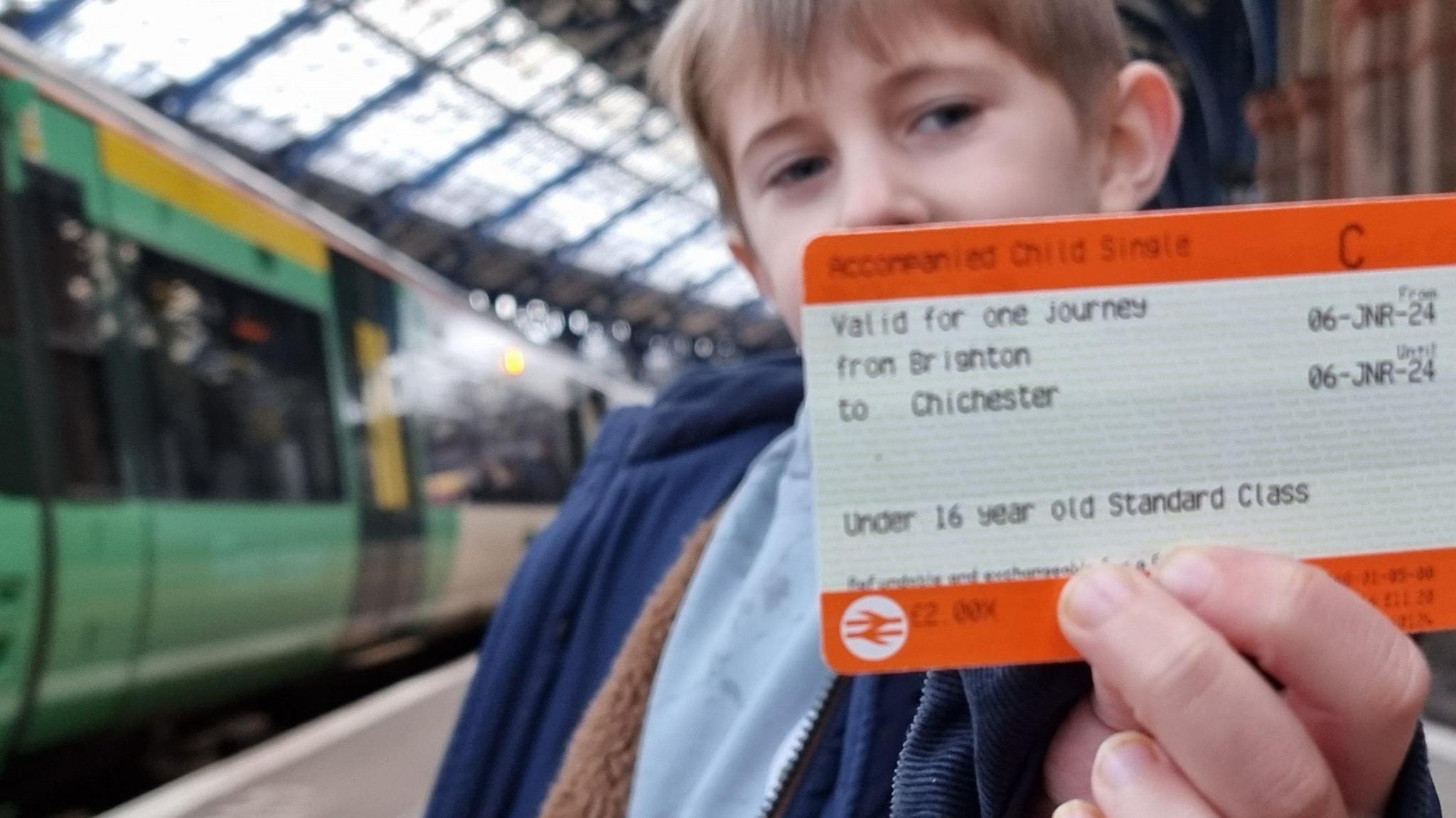 Austin, a boy with short brown hair who is holding an orange train ticket up to the camera. The journey on the ticket says from Brighton to Chichester. He is stood in front of a green train on a platform at a railway station and is wearing a blue coat.