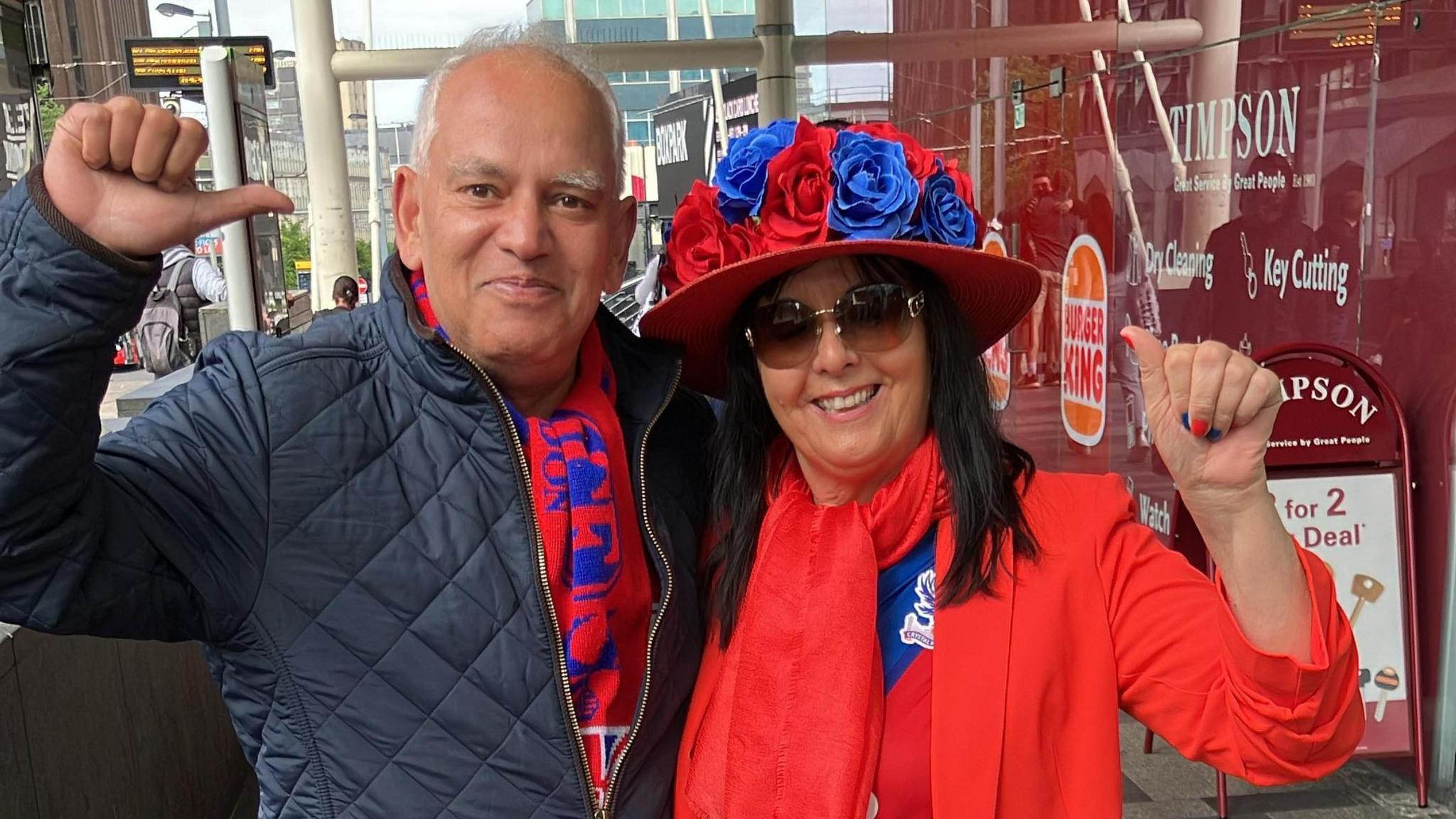 Eugene Borges in a quilted blue jacket and a club scarf. Pamela Gould stands next to him in a red hat covered in red and blue roses. She wears a club t-shirt, red blazer and scarf.