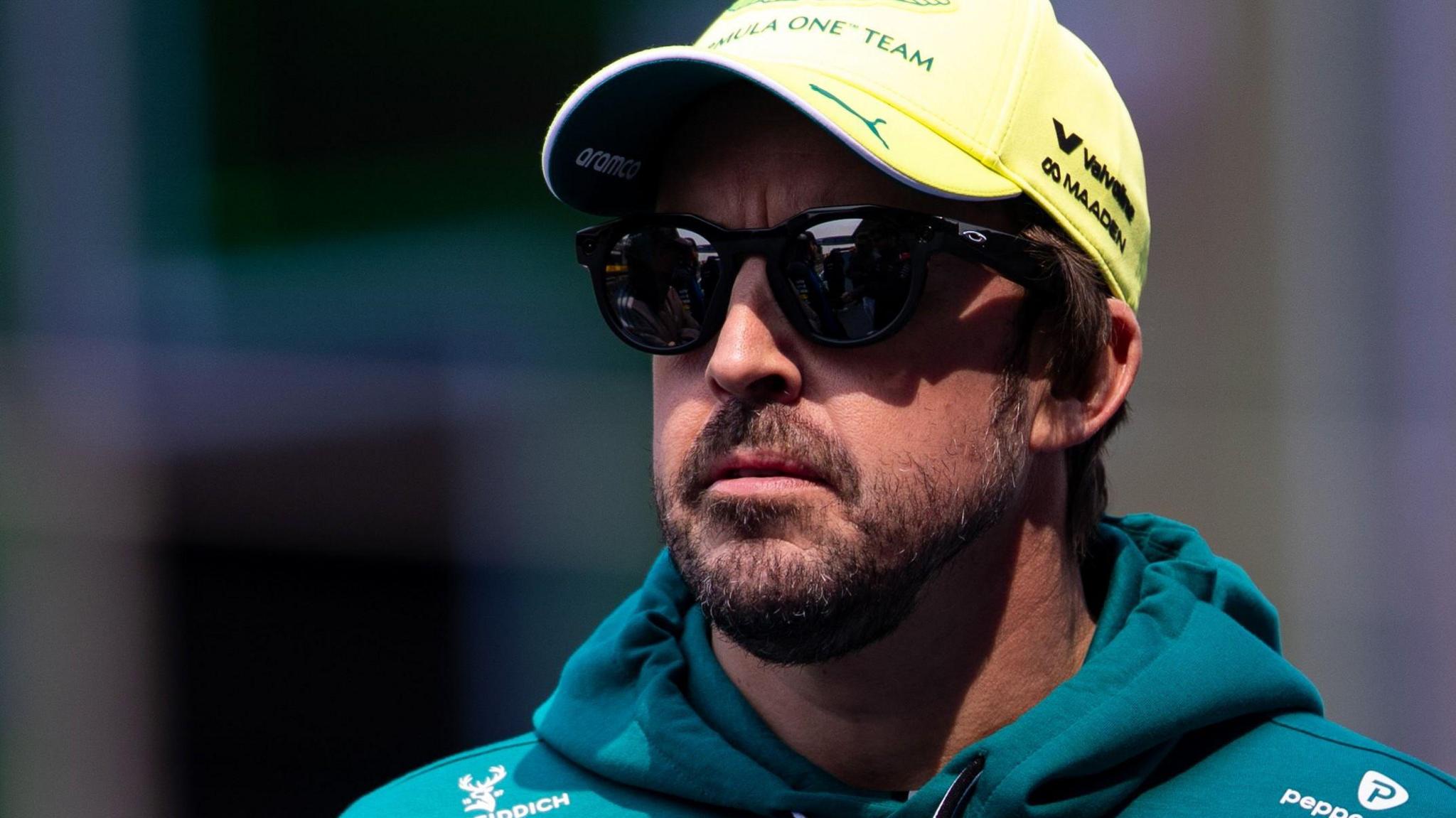 Fernando Alonso wearing sunglasses, an Aston Martin hoodie and a yellow cap
