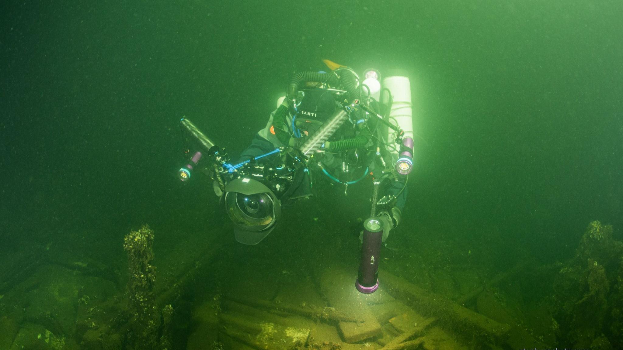 A diver exploring a shipwreck on the bottom of the sea, carrying lots of equipment including a camera and torch