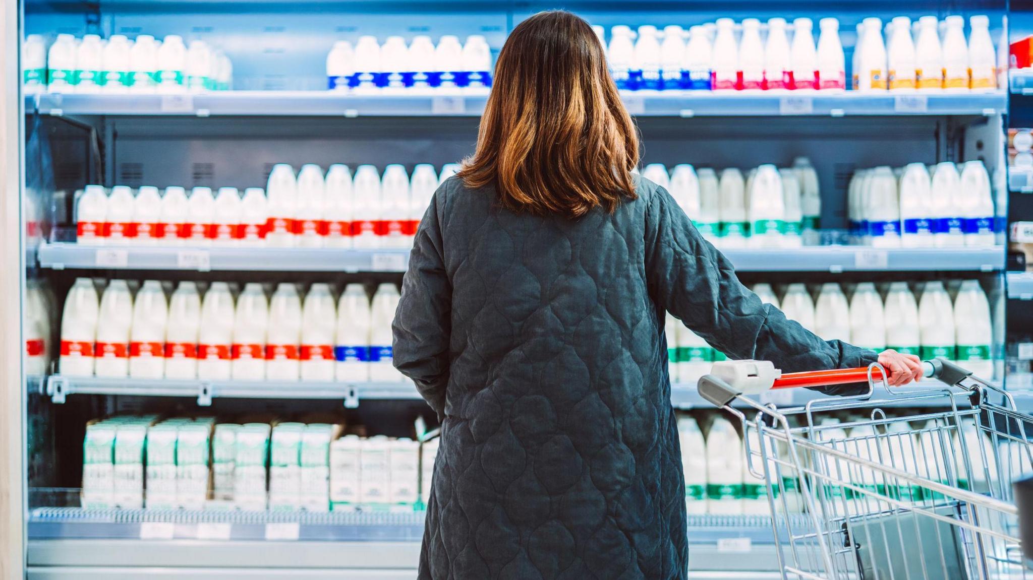 A female shopper, pictured from behind, with longish brown hair and a quilted navy long coat, stares at a fridge in a supermarket full of different types of milk. The brand is not shown.
