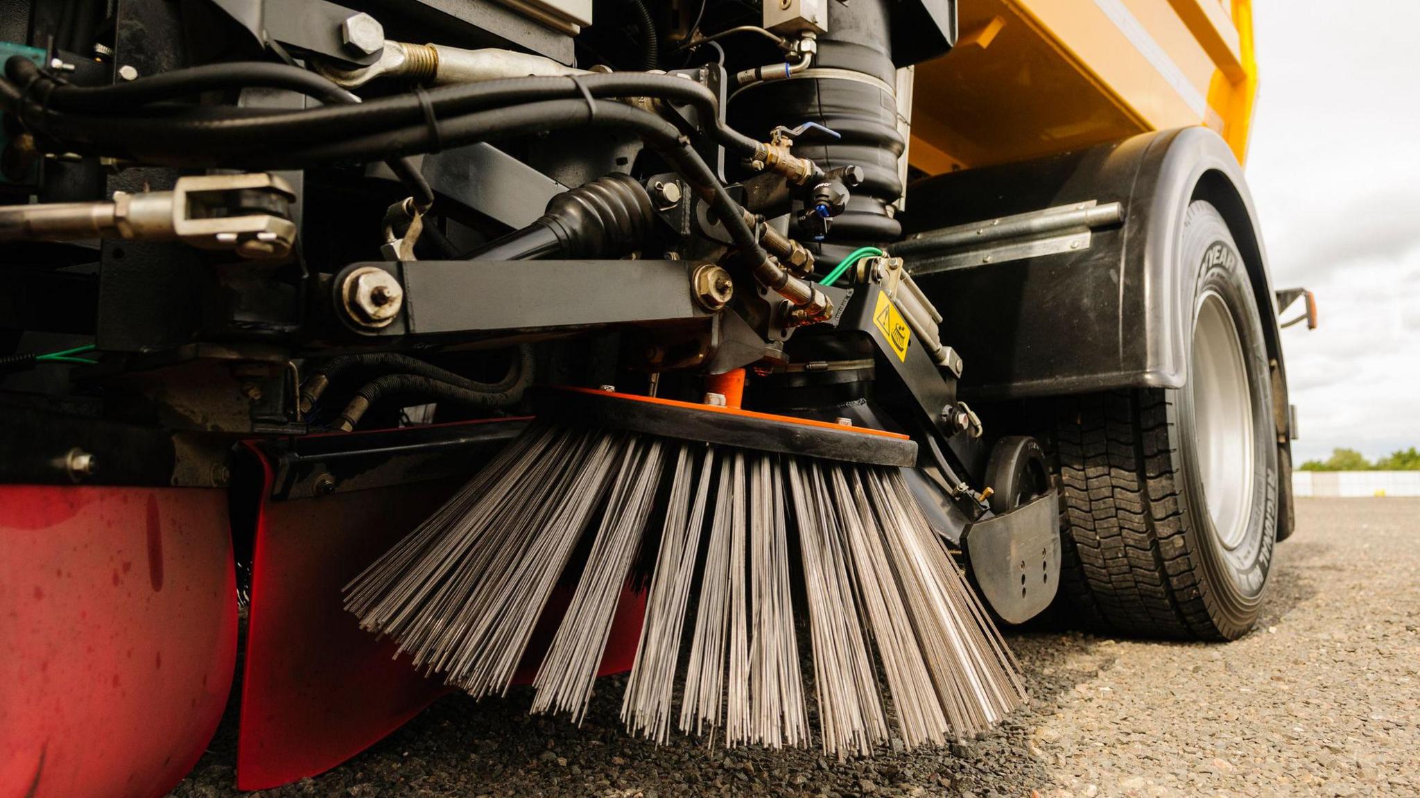 A street sweeper vehicle in a road. Only the sweeping element of the vehicle and its back wheel can be seen