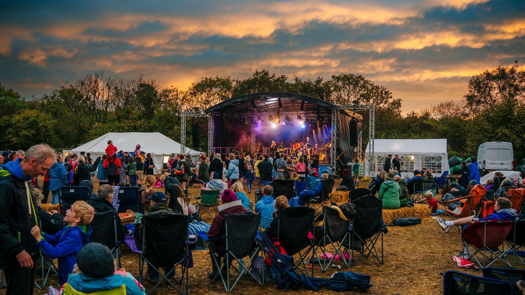 A view of a festival stage from the crowd. People are sitting on camping chairs. The sky is cloudy with the sunset colouring it orange.