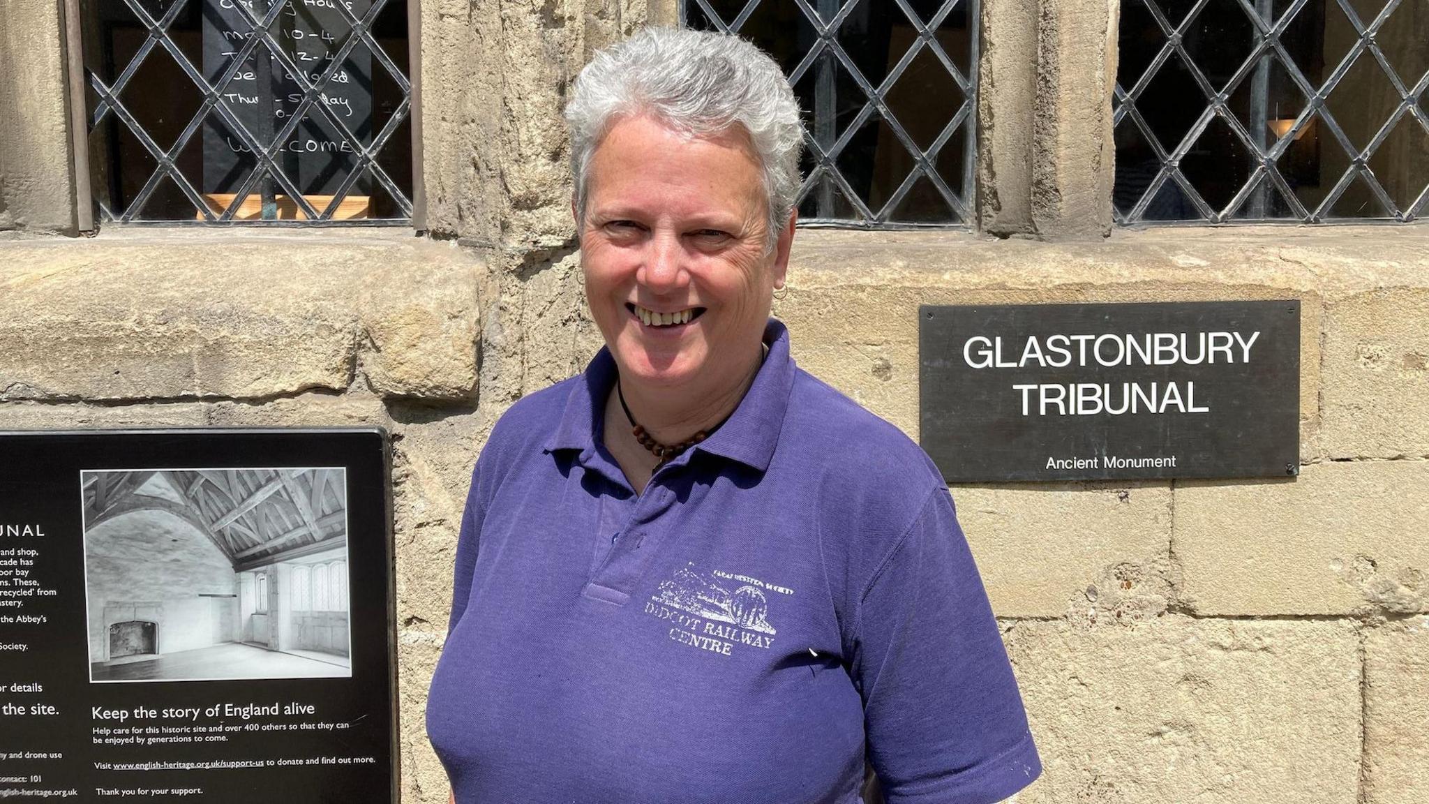 Wearing a purple polo shirt, Pauline is smiling at the camera, and standing next to a sign saying "Glastonbury Tribunal".