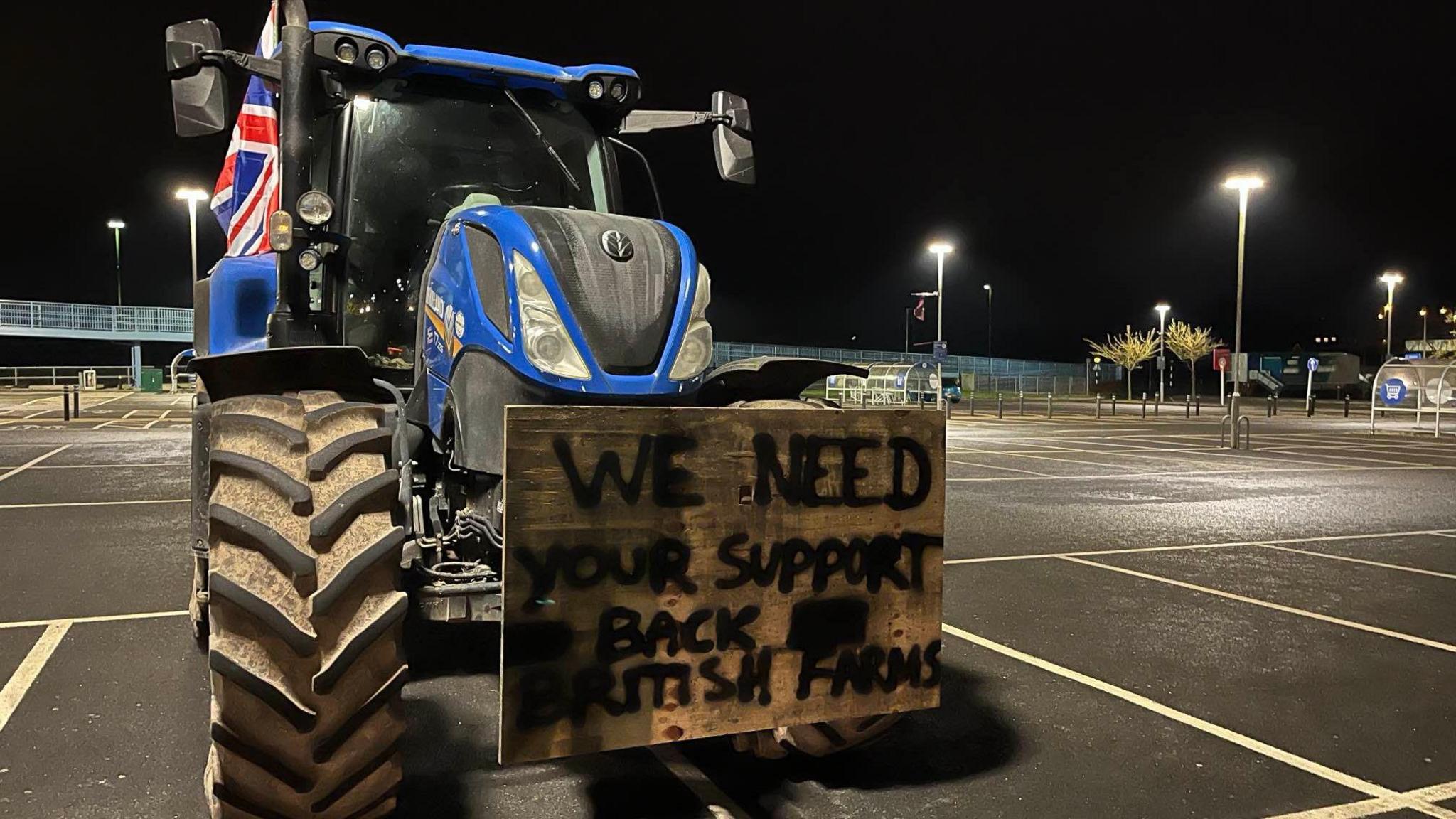 A blue tractor is parked in a supermarket car park and has a wooden sign on its hood reading: "We need your support - back British farms."