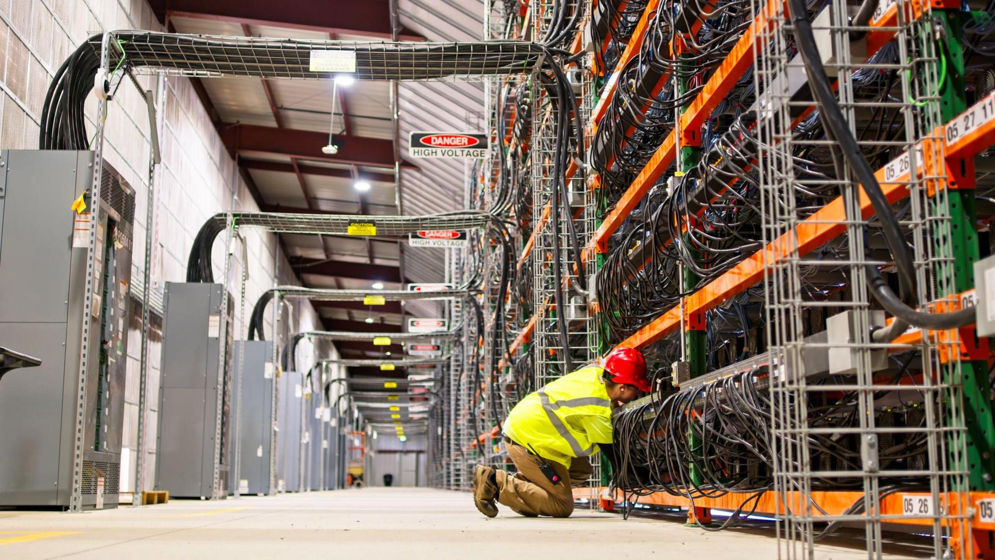 A technician in a high-vis jacket and hard hat kneels on the floor of a warehouse, fixing computer wiring on a series of racks towering above them.