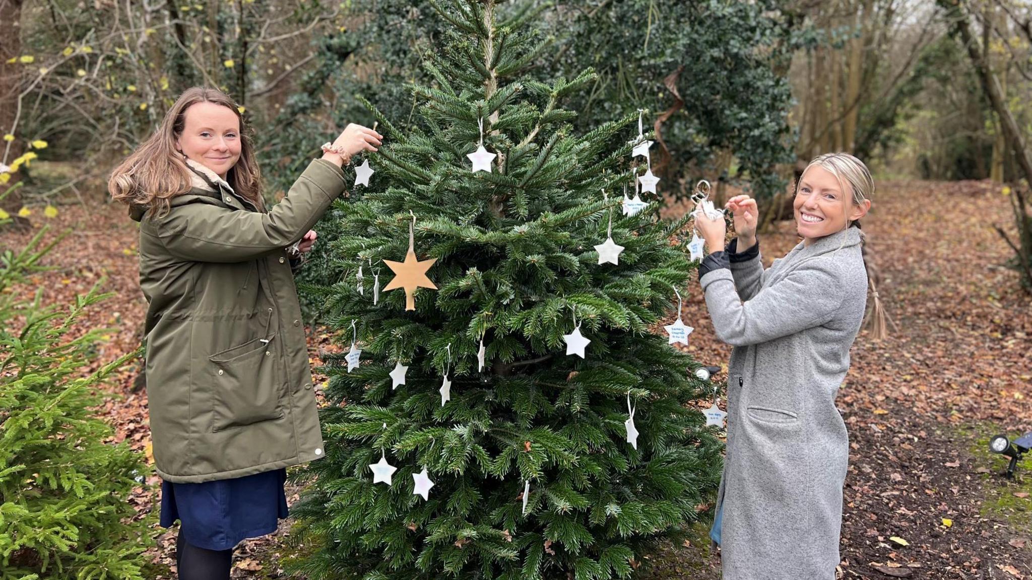 Two women standing in woodlands and putting Christmas decorations on a tree in the shape of white starts. They are both wearing coats and smiling at the camera.