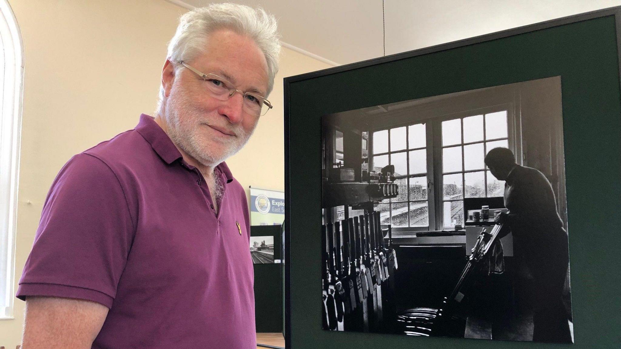 David Pearce smiles at the camera while standing next to a black and white image of a man standing in a railway operator room. David has white grey hair and wears a burgundy polo T-shirt.