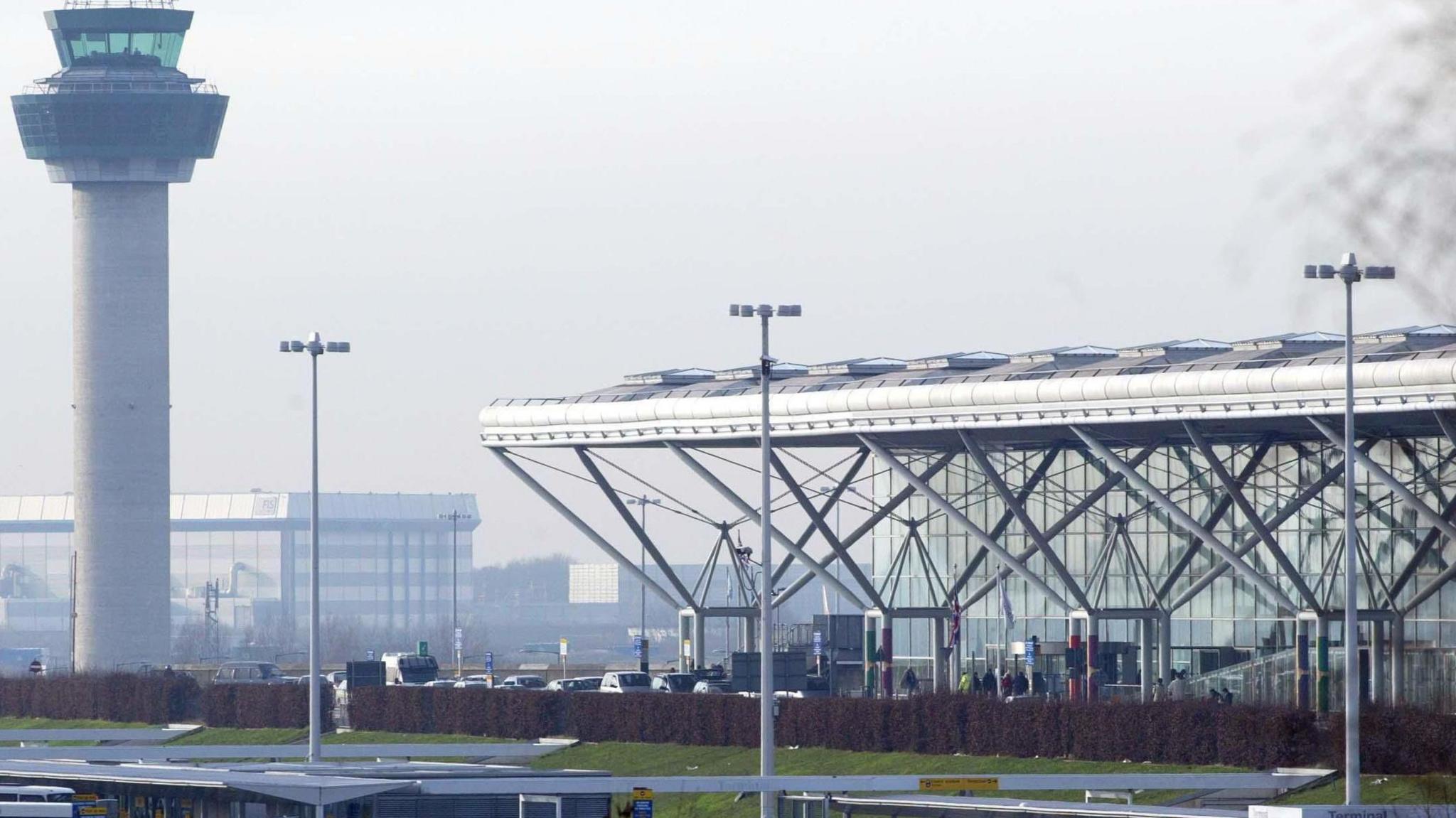 London Stansted Airport on a cloudy day. The airport control tower is to the left and the main terminal buiding, which has a glass facade and a white roof supported by grey pillars.