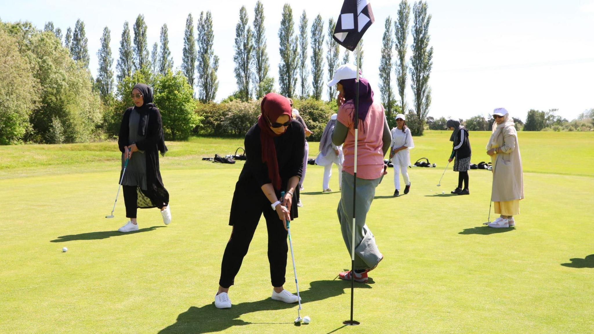 A group of women on a golf course. Some of the women are dressed in traditional Islamic clothing such as long, loose-fitting tunics and hijabs. Others are wearing caps and sportswear.