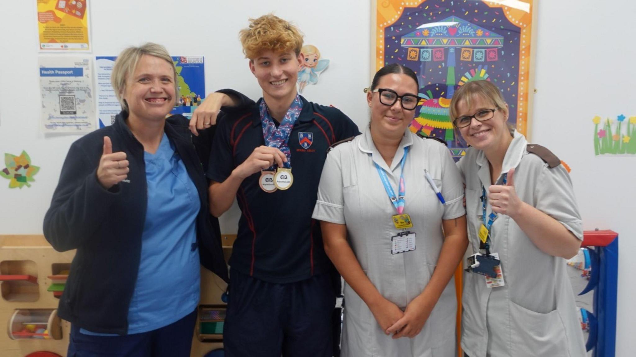 A young man with short curly hair and wearing a dark top stands in a line with three women in medical scrubs. Two of the women are giving a "thumbs up" gesture to the camera and all four people are smiling