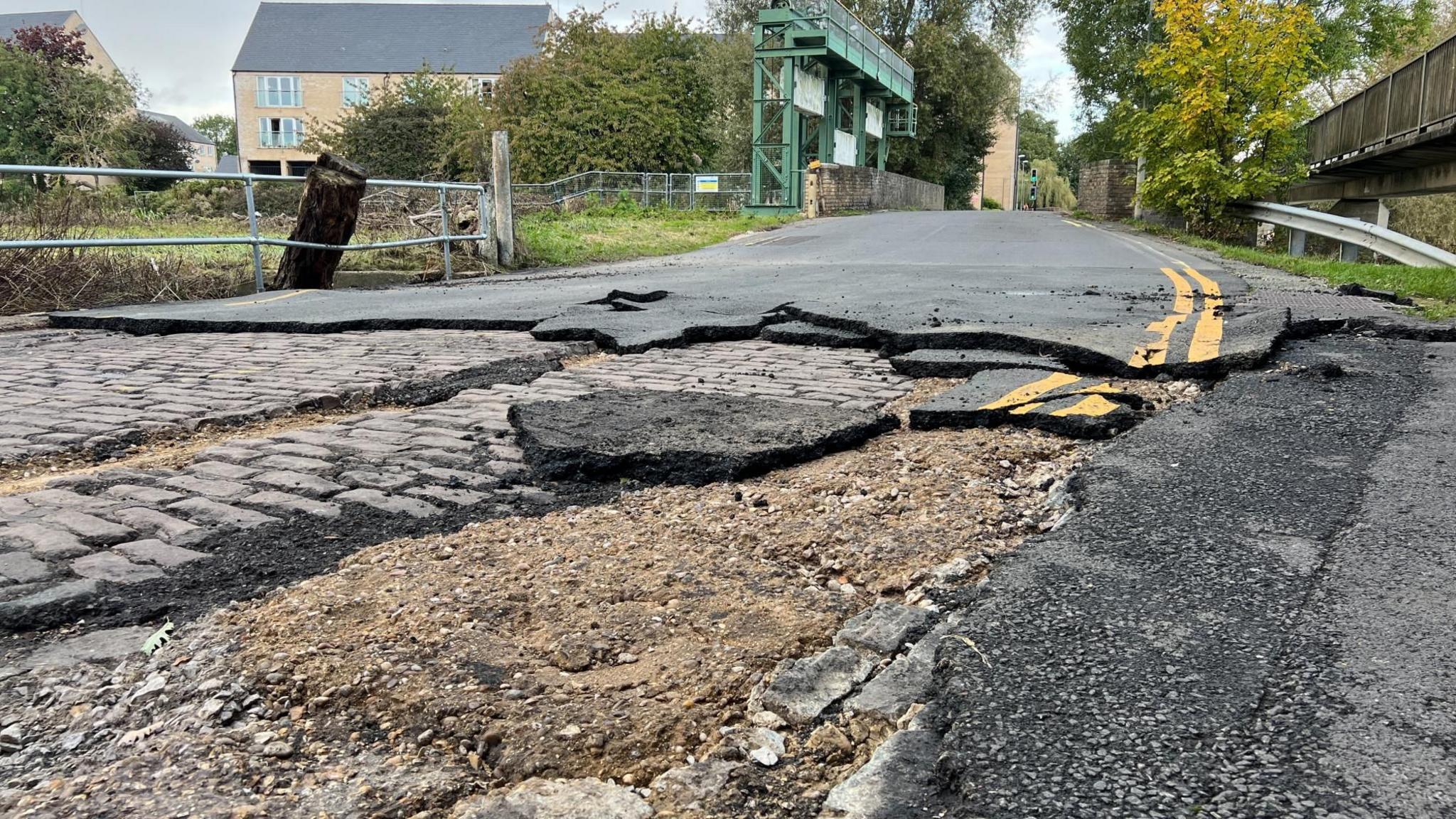 A ripped up road surface revealing cobbles underneath, with the bridge and weir over the River Great Ouse between St Neots and Little Paxton