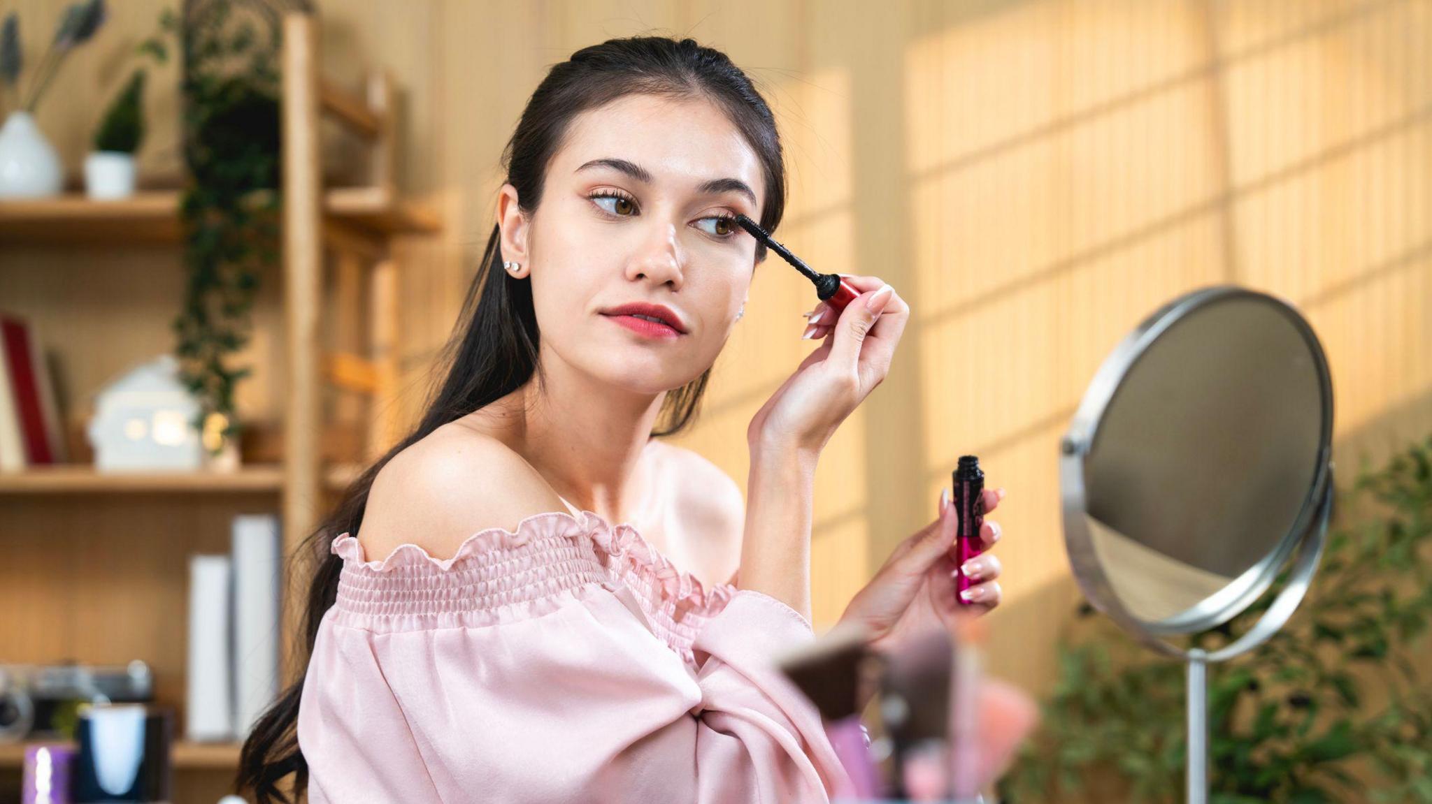 Woman with long dark hair, wearing a pink off-the-shoulder top looking in a small mirror while she applies mascara