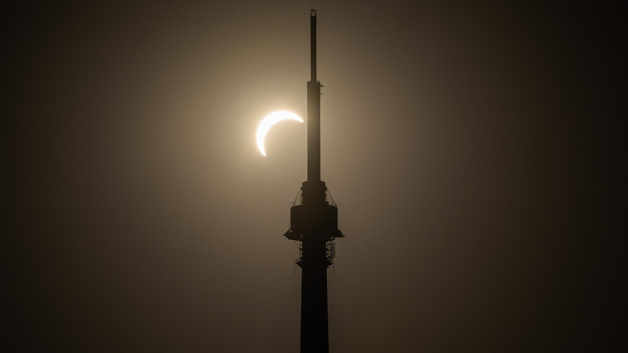 A partially covered sun lights up the dark sky. It can be seen from the silhouette of a large tower.