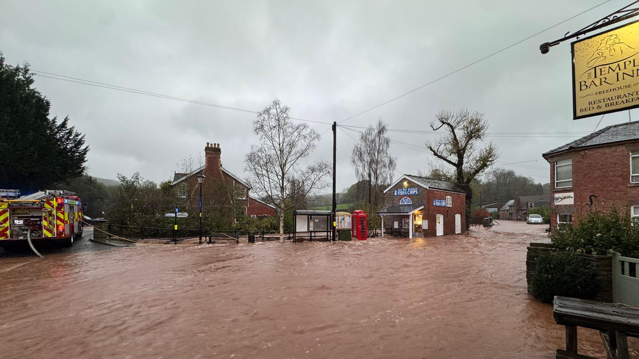 Dark orange floodwater running through a village. The water has covered the road and is sweeping through the street. There is a pub on the right and a bus stop, phone box and fish and chip shop that are all flooded. A fire engine is on the left trying to pump out the water.