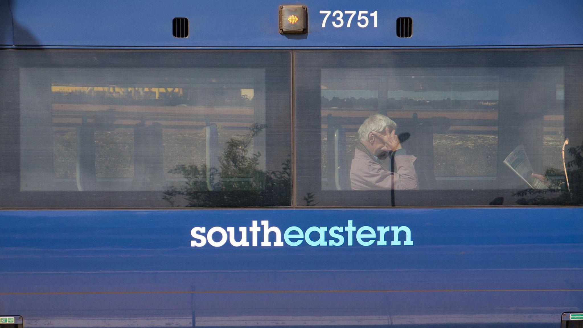 The side of a blue Southeastern Railway carriage. Behind one of the two windows, commuters sit waiting for the train to continue its journey.