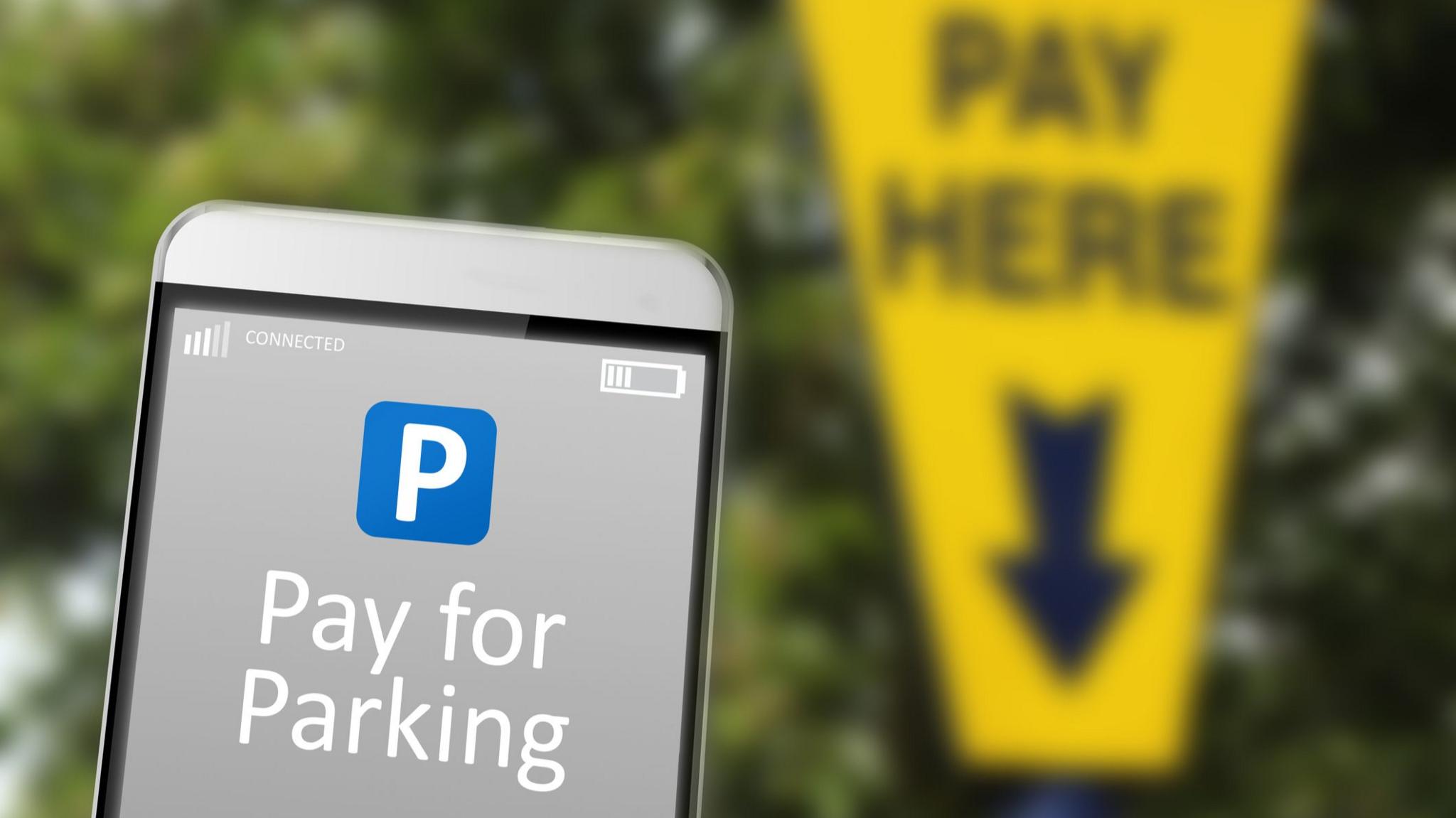 A generic image of a car park app on a phone in the foreground with a pay here parking meter sign in the background