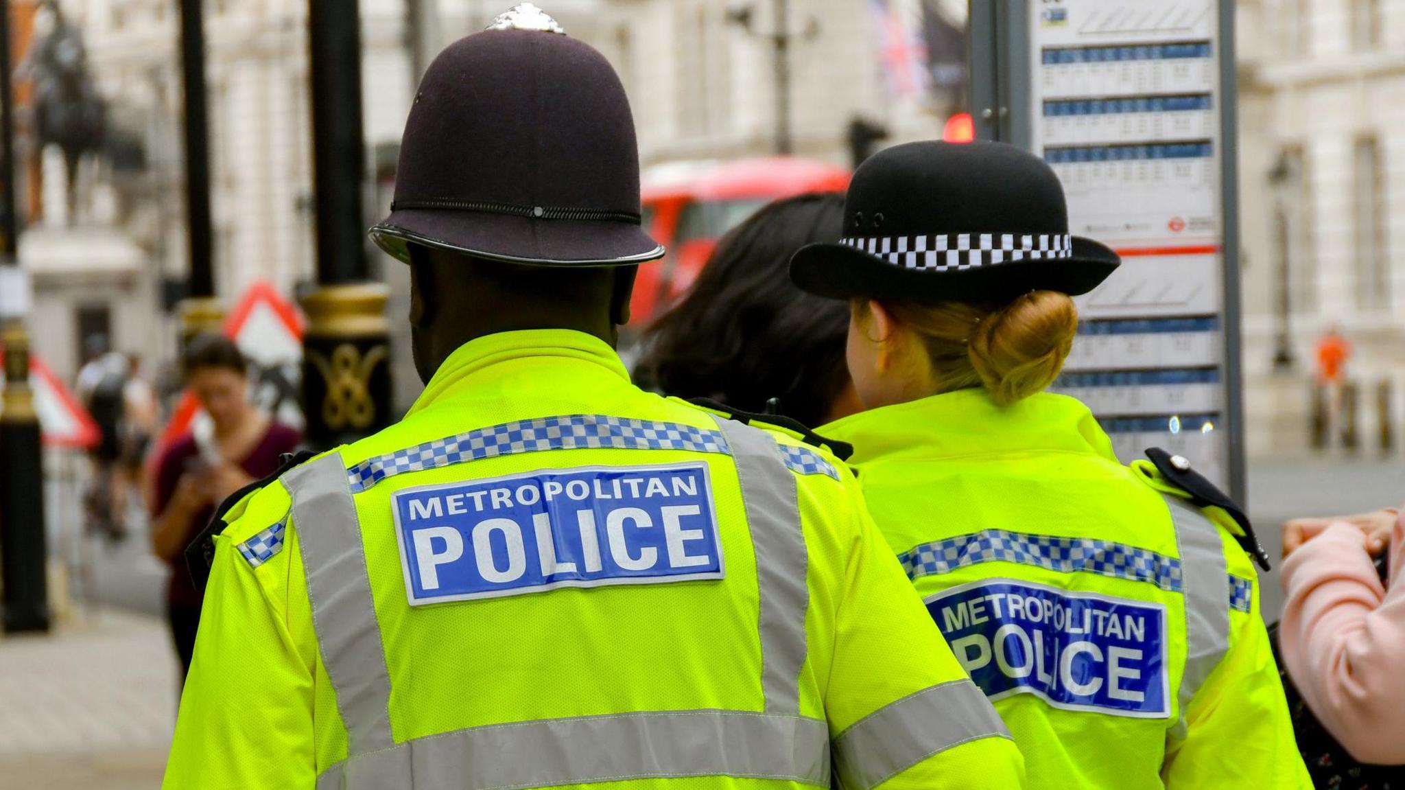 The back of two Met police officers, one male, one female. They are wearing yellow high-visibility jackets with "Metropolitan Police" written in white on a blue background.