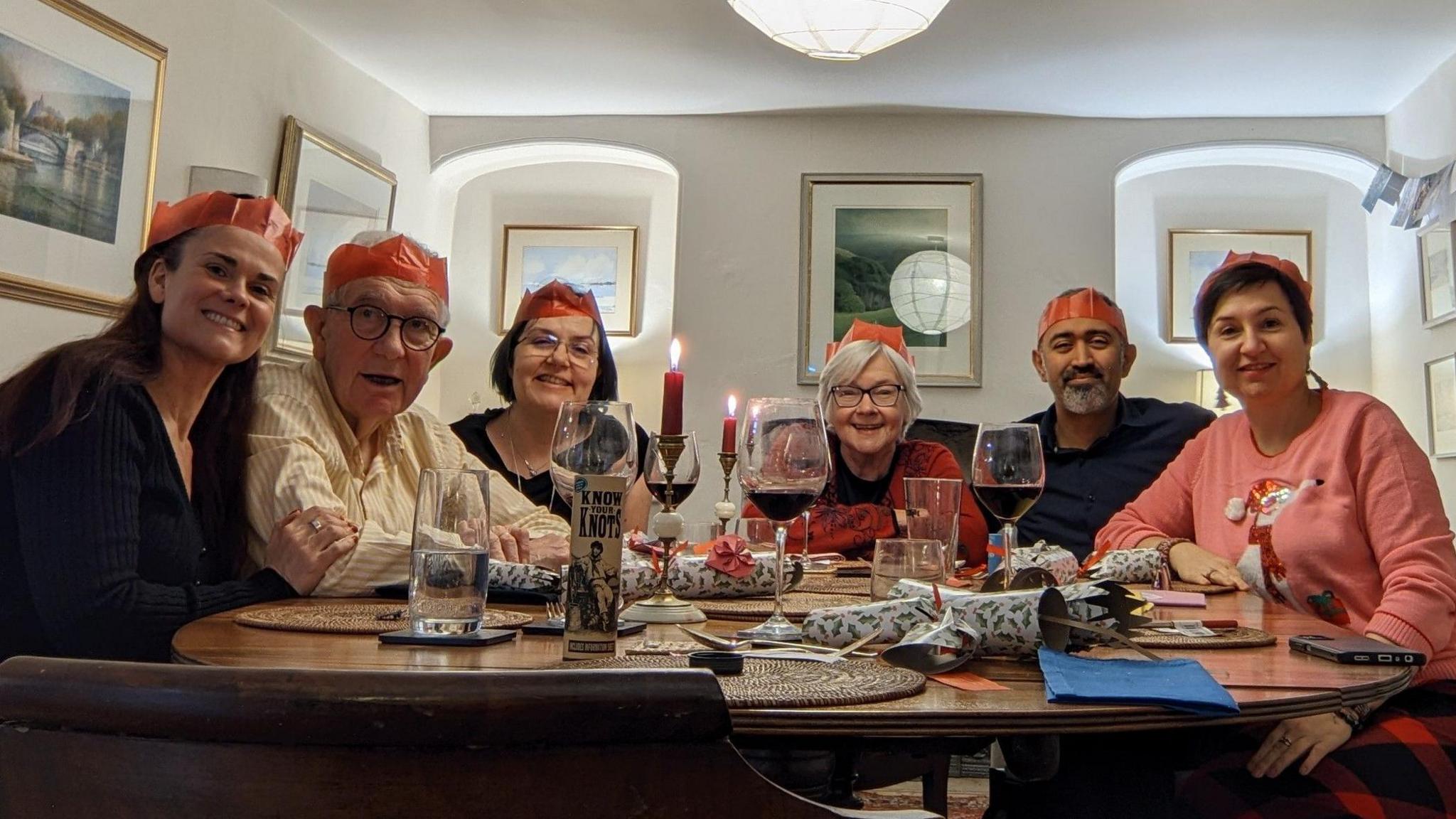 Six people sitting around a table for dinner wearing orange Christmas cracker crowns.