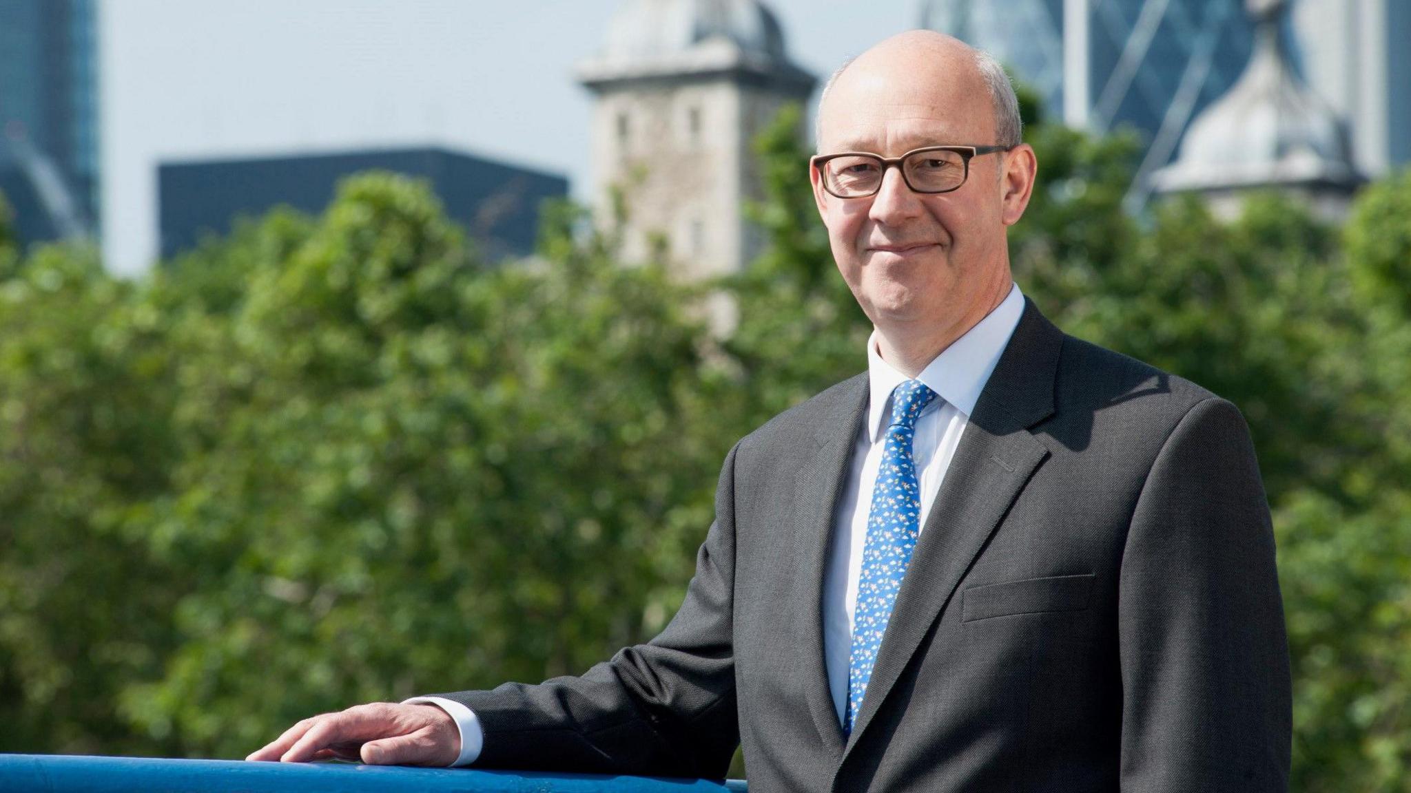 A file image of Simon Birkett, a man wearing glasses and a grey suit with a blue tie, with his arm resting on a blue railing. Trees, buildings and skyscrapers can be seen in the distance