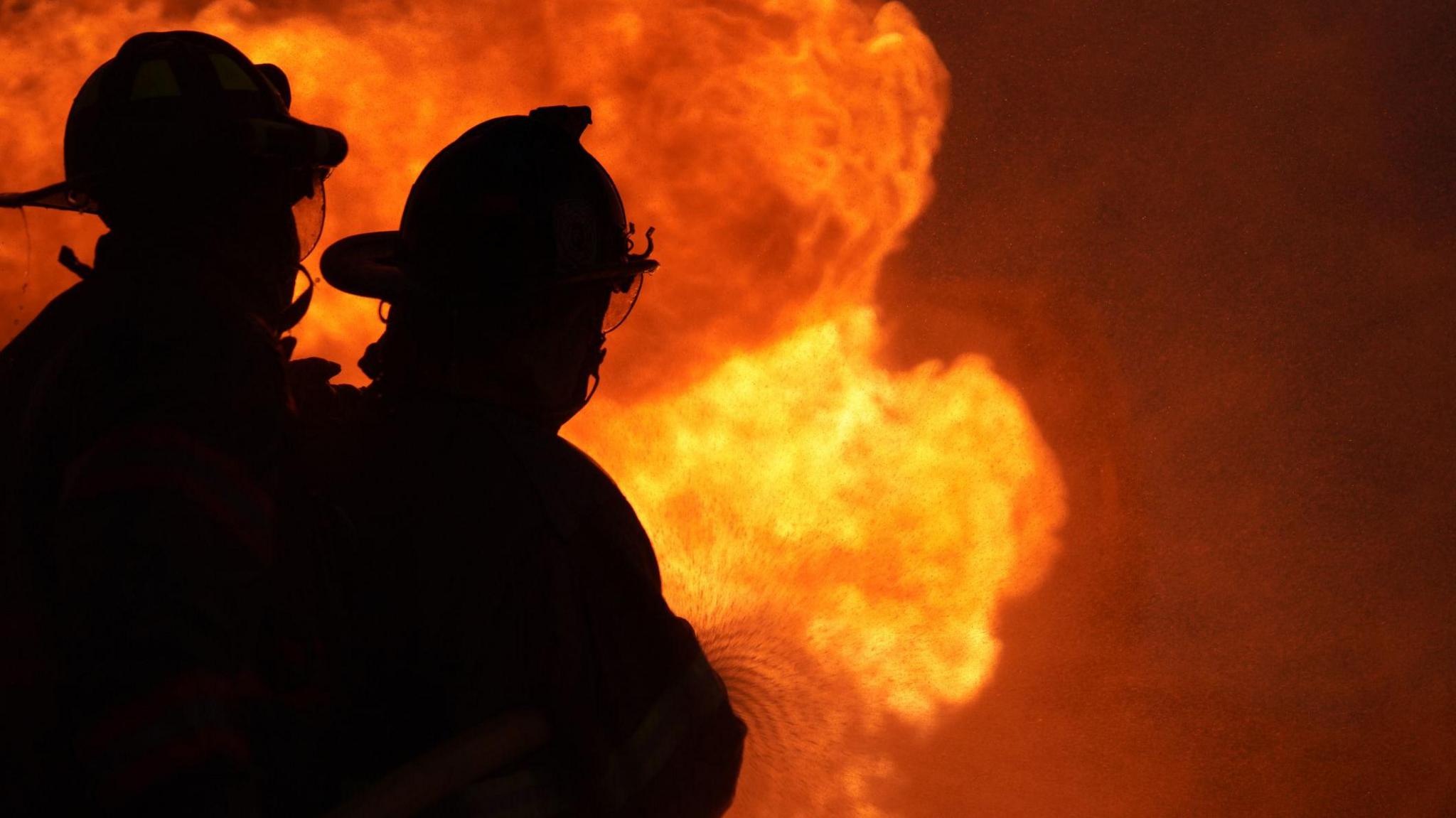 Stock image of two firefighters, shown in silhouette, battling a blaze. The flames are bright orange and yellow.