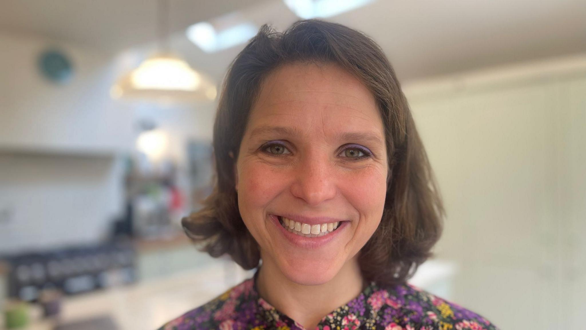 It's a close up photo of a short, brown-haired Eliza smiling at the camera and wearing a floral shirt. The background is blurred. 