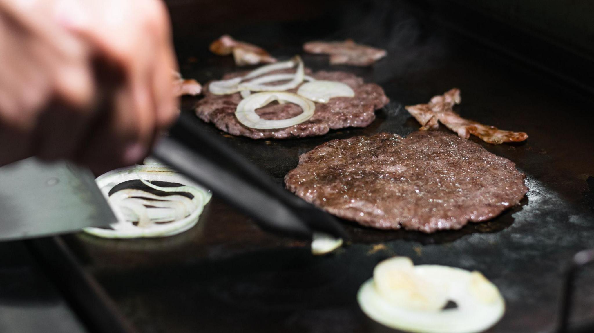 A hand can be seen flipping a burger on a grill, there are also onions and bacon being cooked on the same grill.
