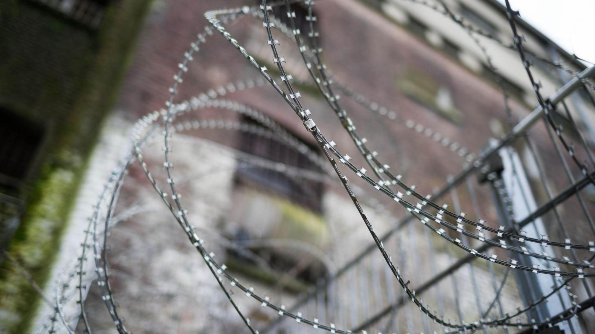 A blurry picture of barbed wire fencing at a prison. We can see a big and intimidating building block behind. The wire is razor sharp.