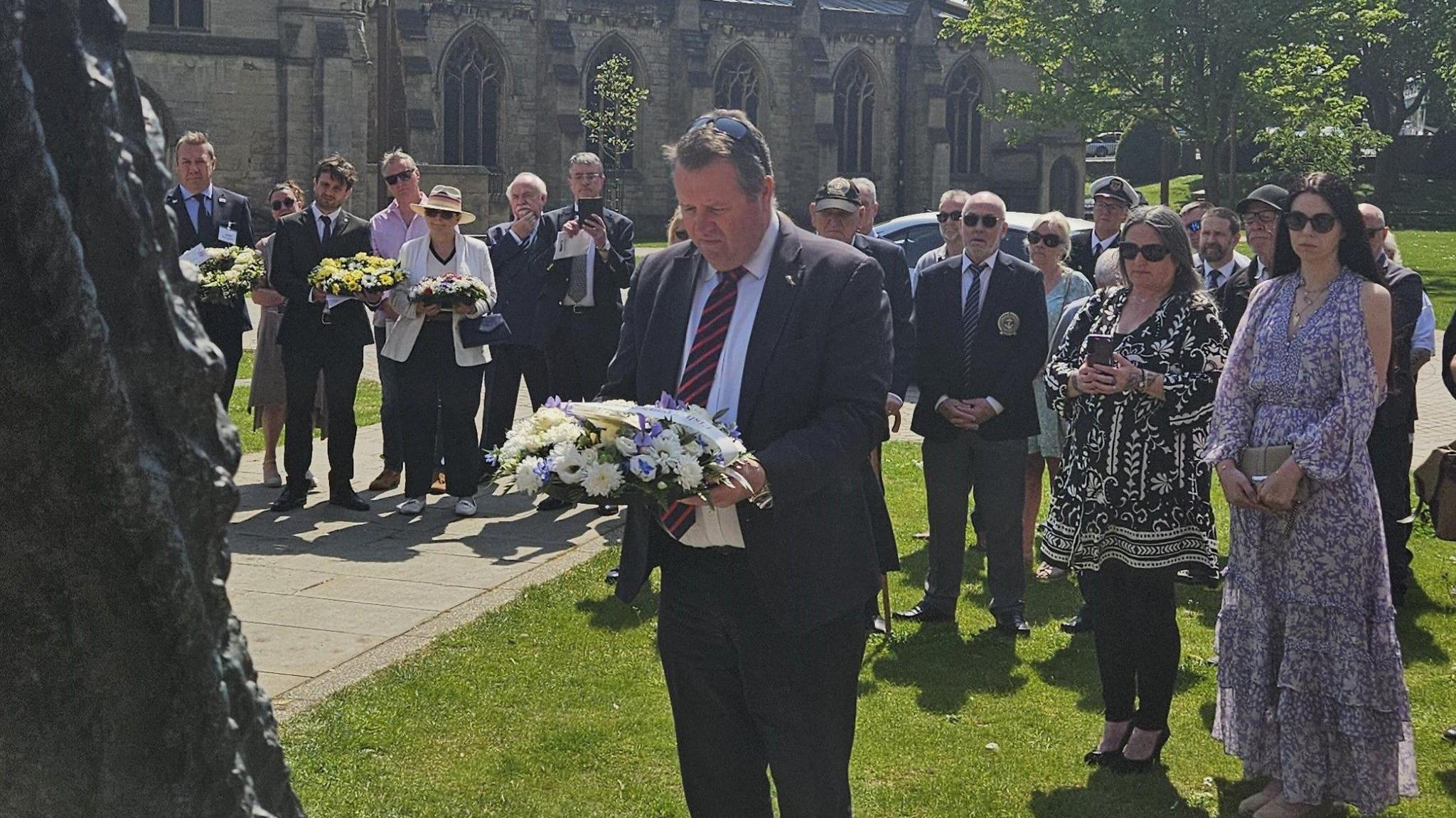 A small crowd of smartly dressed people attend a service outside a church on a sunny day. In the foreground, the former Fisheries Minister Sir Mark Spencer lays a wreath of flowers at a memorial.