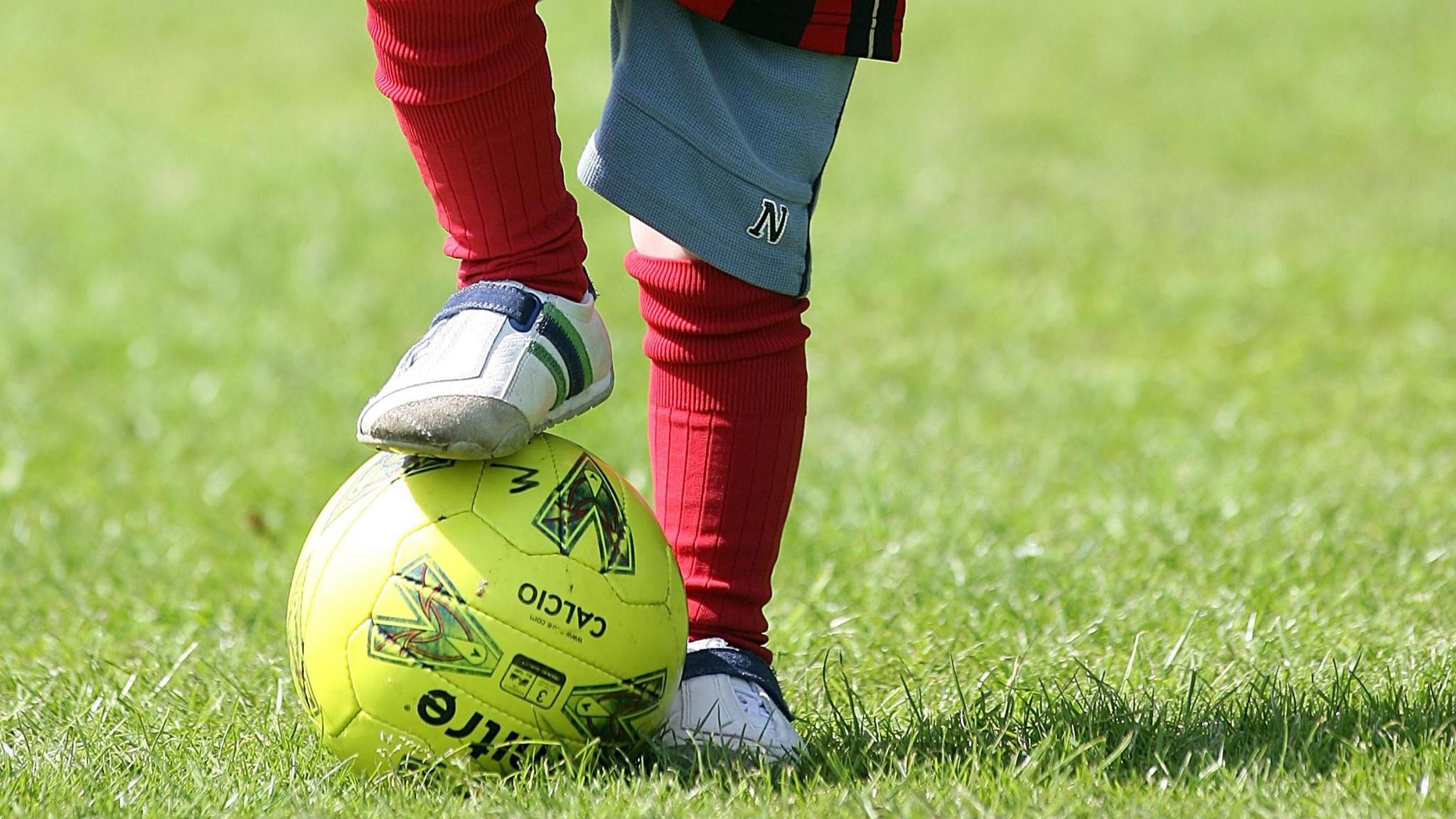 A child is balancing a yellow football on the grass with his foot on top of it. You cannot see the child's face. They are wearing red socks and white trainers