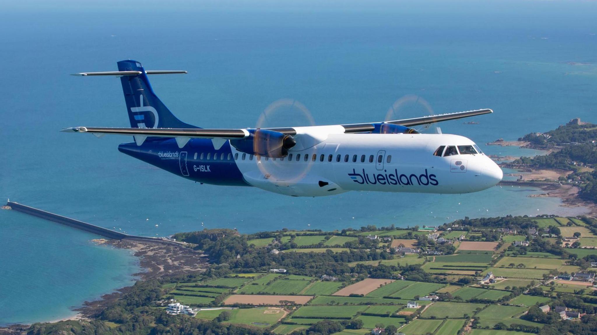 A file photo of a plane in the sky flying over land. There is green land on the right and sea on the left.