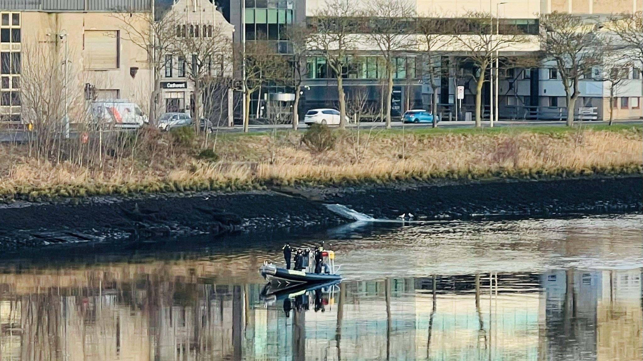 A small police boat on a river, with cars and buildings in the background.