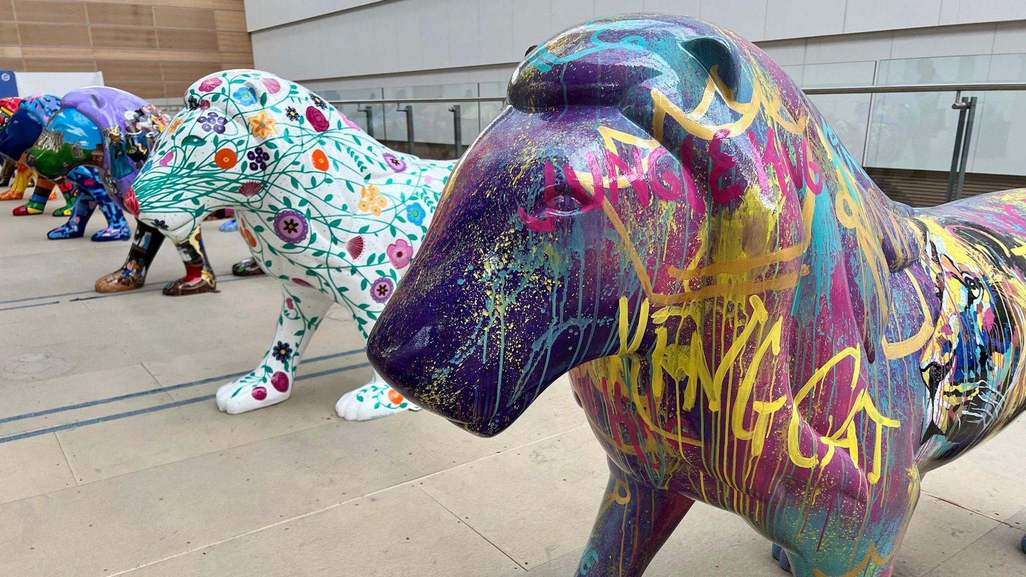 A row of the painted lions inside a large inside space. They are all brightly coloured, and the one closest to the camera is purple and covered with a graffiti pattern. The one next to it is white, covered with green leaves and multicoloured flowers.