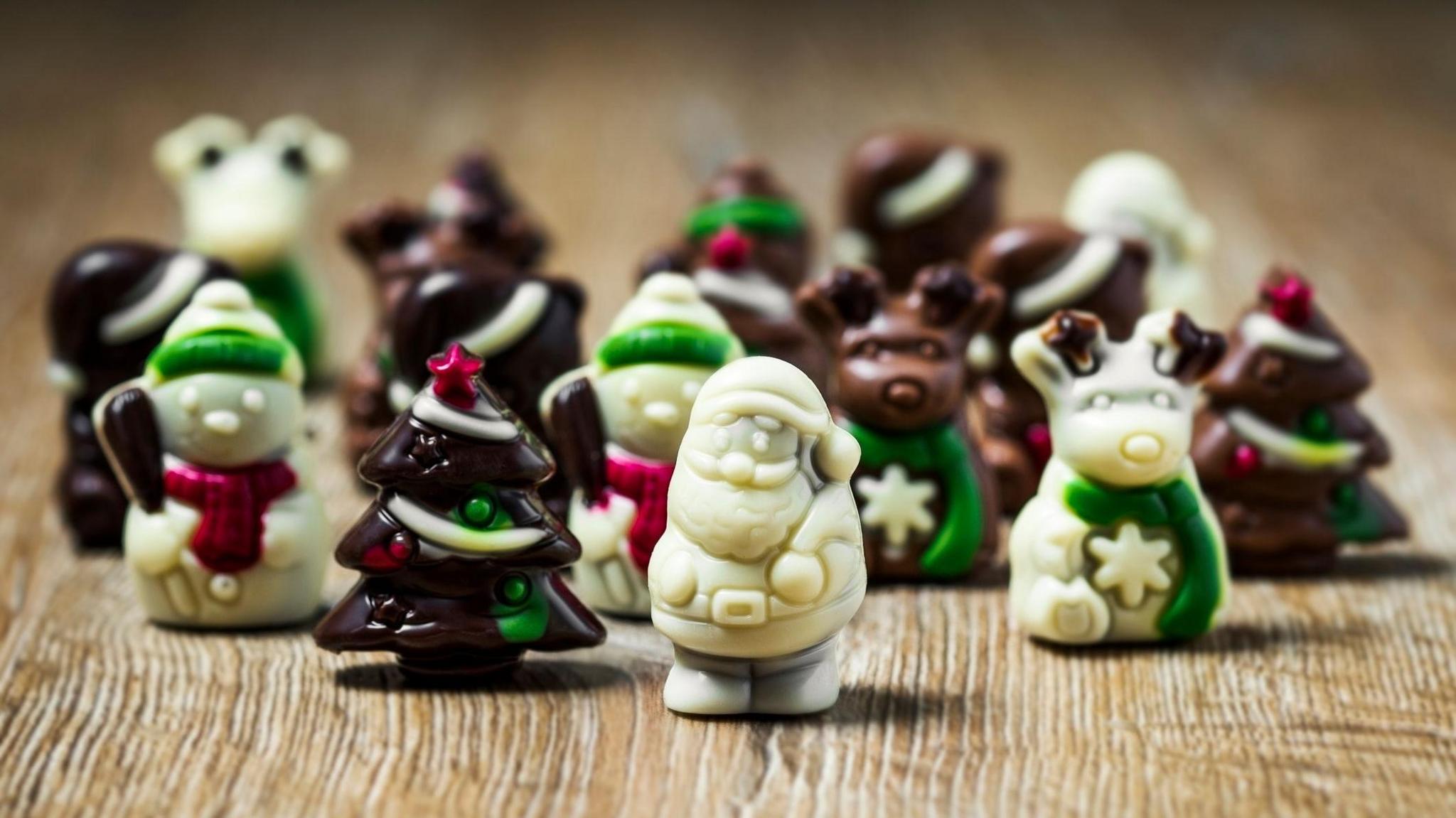 A mixture of chocolates in different Christmas shapes are standing upright on a wooden table. There are white chocolate santas, reindeer and snowmen as well as milk and dark chocolate christmas trees and reindeer. 