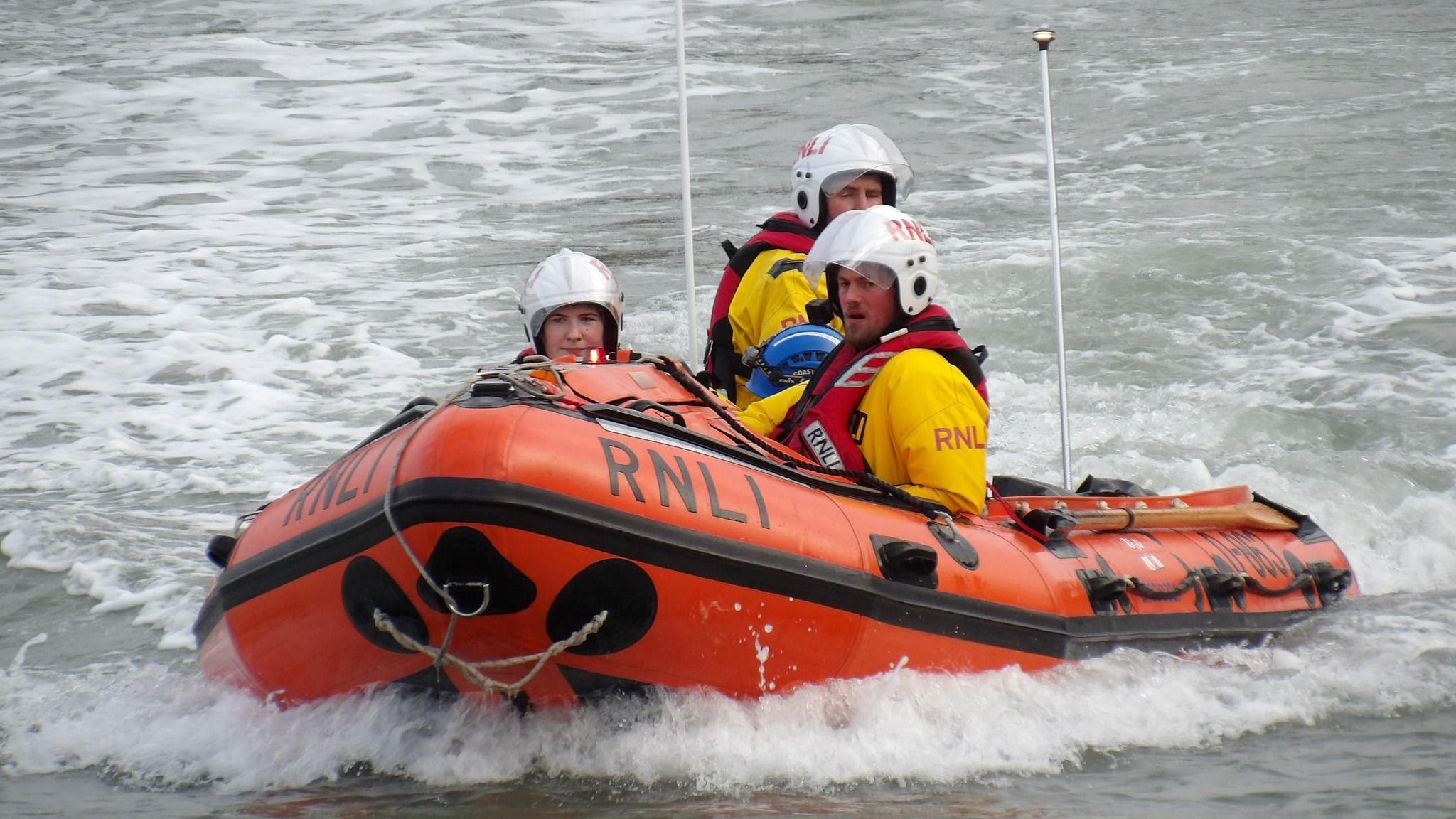 RNLI Ilfracombe's inshore lifeboat with three crew members aboard who are in uniform and wearing lifebelts and helmets. The RIB reads RNLI on its bow.