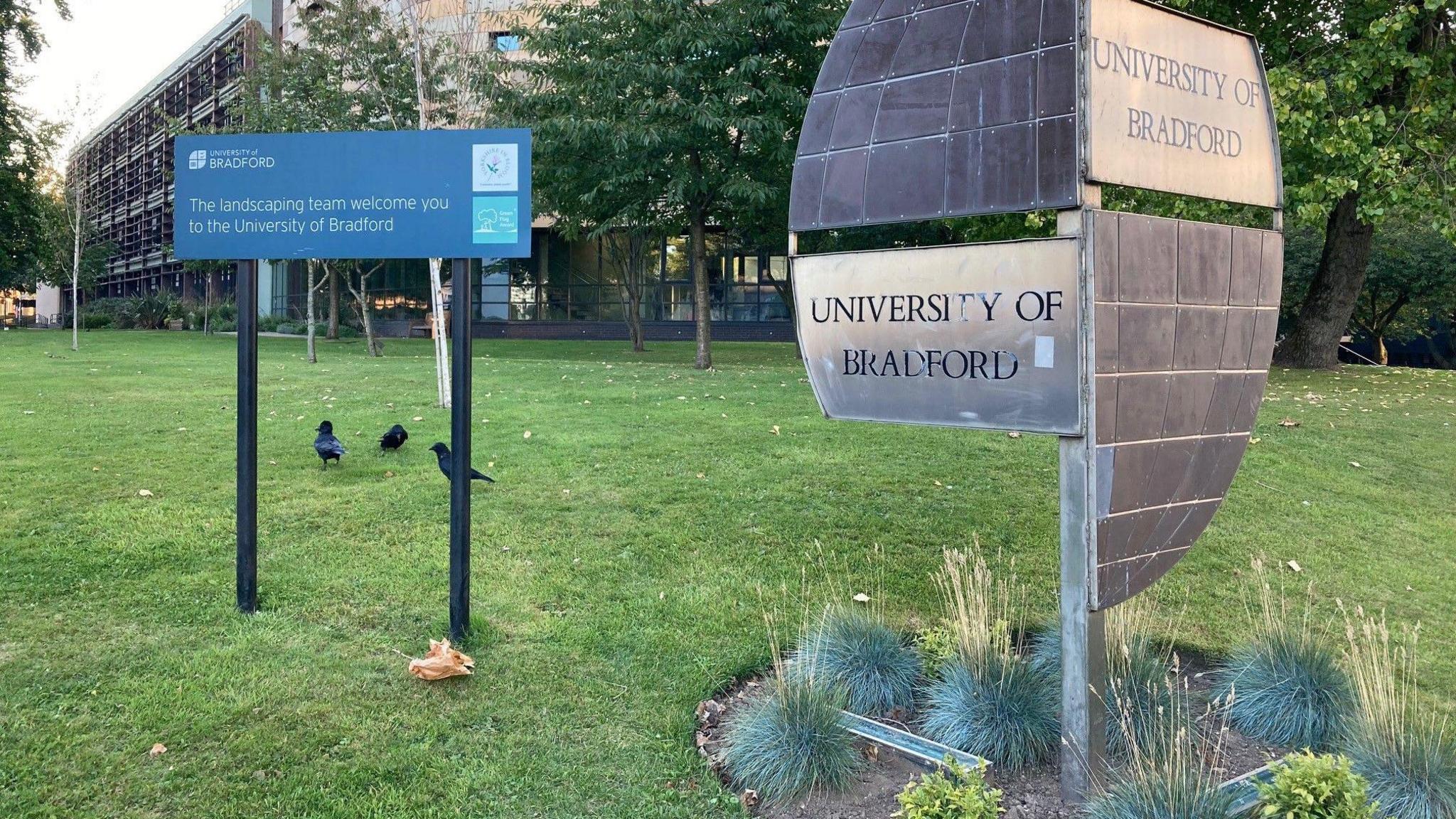 A blue sign on a grass verge on the campus welcoming visitors to the University of Bradford alongside a metal, sculptural-type sign spelling out the University of Bradford