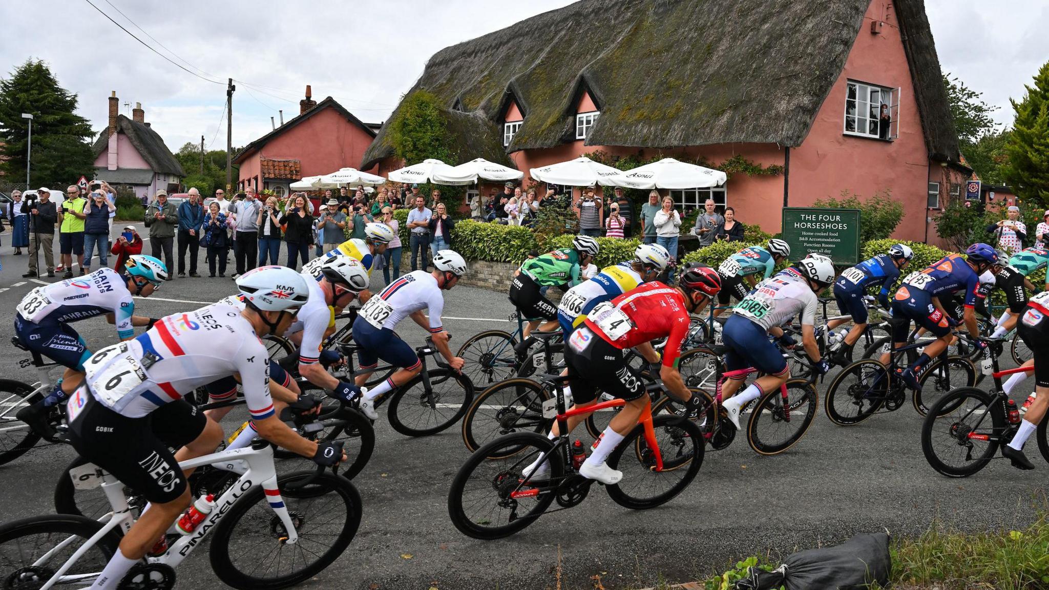 Cyclists race around a corner past a thatched roof pub. Spectators stand at the side of the road to watch on and cheer the racers.