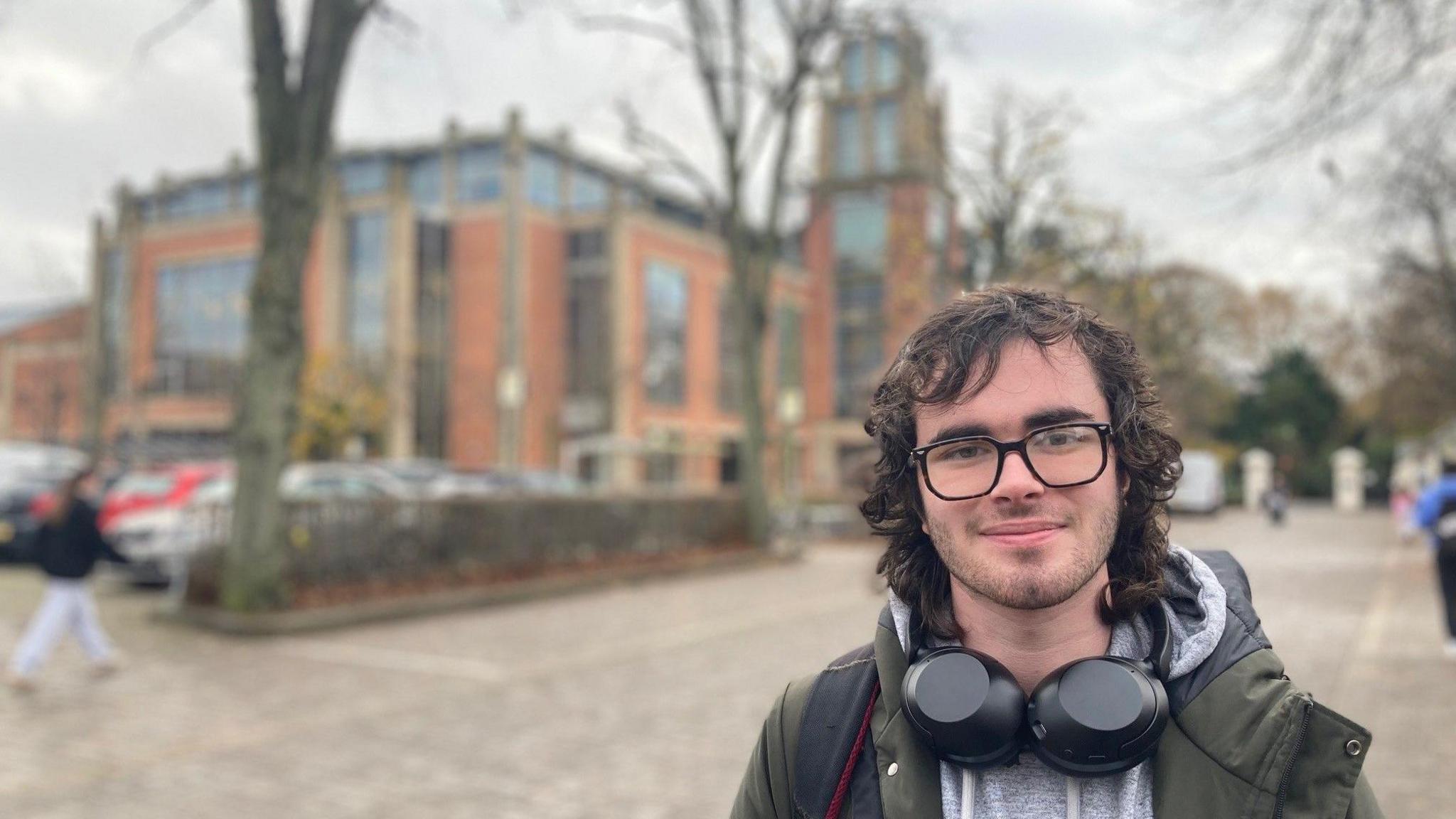 A person looking at the camera. They are wearing a grey hoodie and a dark green jacket. They have large black headphones around their neck and glasses on their face. They have long dark hair. Behind them, there is a large red-brick building, as well as a number of people walking. There are also some parked cars.