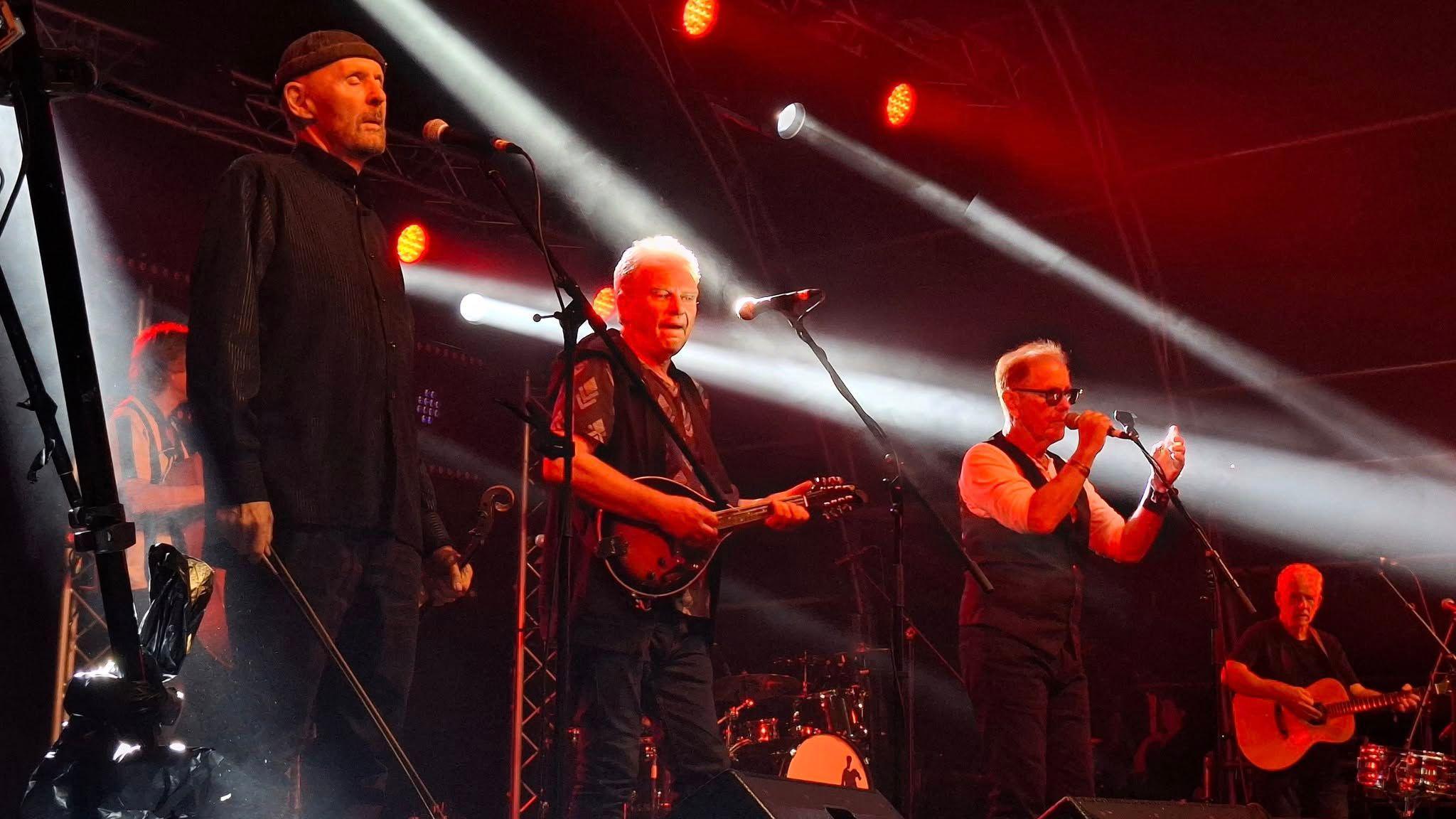 Three older men performing on a dark stage that is lit with a few red lights and white spotlights. They are all standing behind microphones.