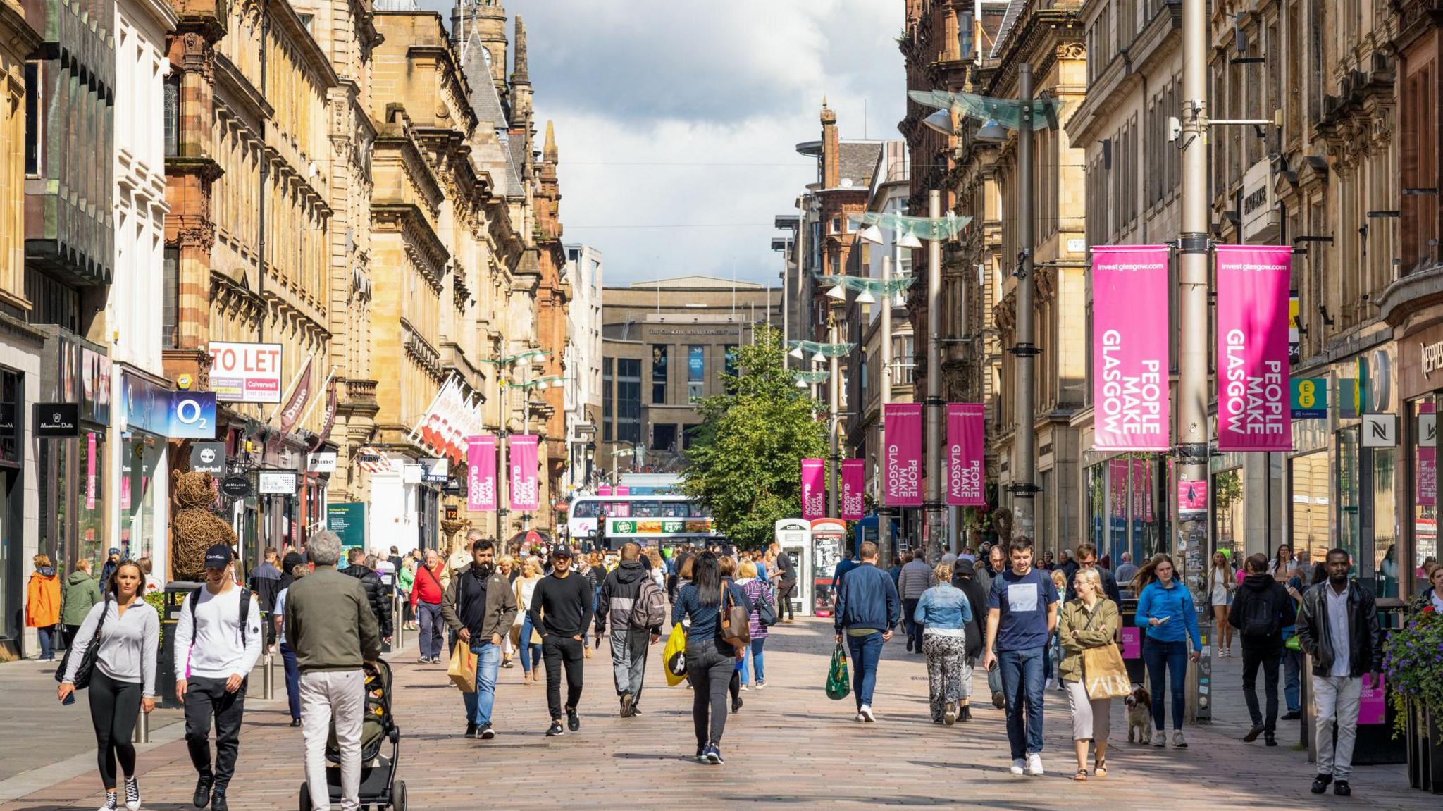 A stock image of Glasgow's Buchanan Street. There are crowds of shoppers walking in both directions. On the lampposts there are banners which say 'People make Glasgow' on them. Multiple shops are visible on the left and the right of the image.