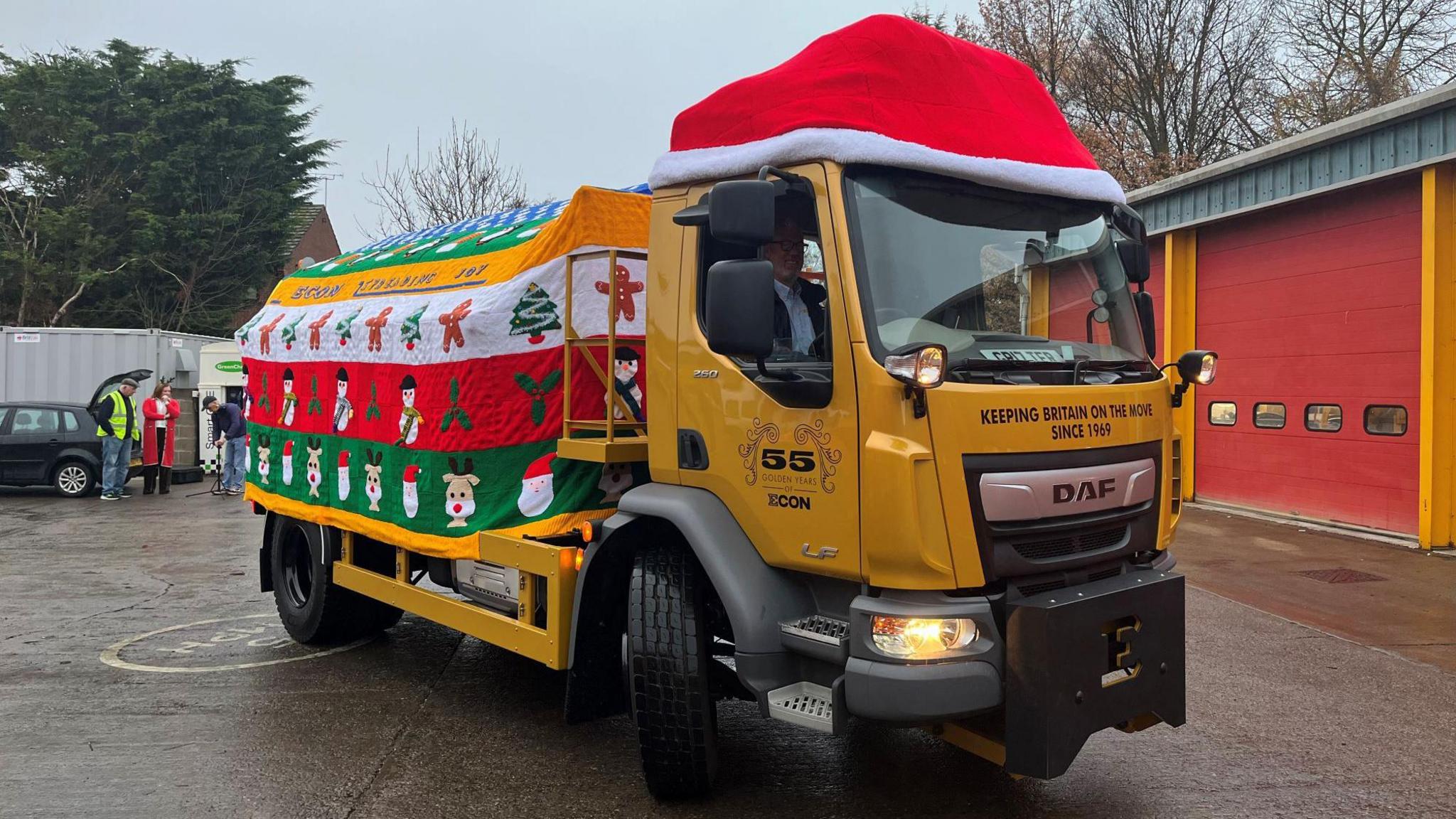 A yellow gritter covered in a "knitted jumper" and a Santa hat. Pictures of gingerbread men, reindeer and Santa make up the design.