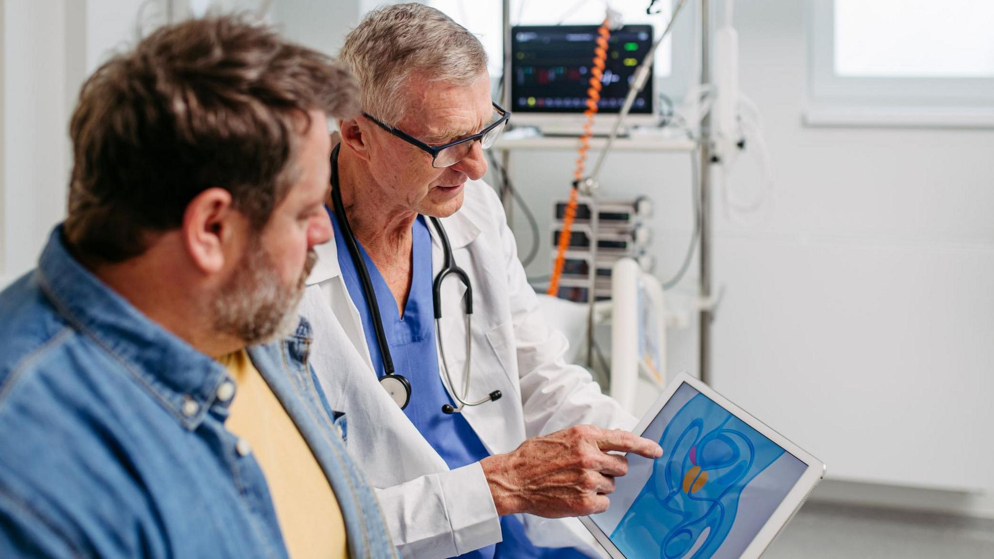 A man with short brown hair and a light yellow T-shirt under an open denim shirt sits on the left of the image, watching as a doctor demonstrates the location of the prostate on a tablet. The doctor, wearing blue scrubs, a white medical coat, black-rimmed glasses and a stethoscope, holds the tablet while explaining the image.